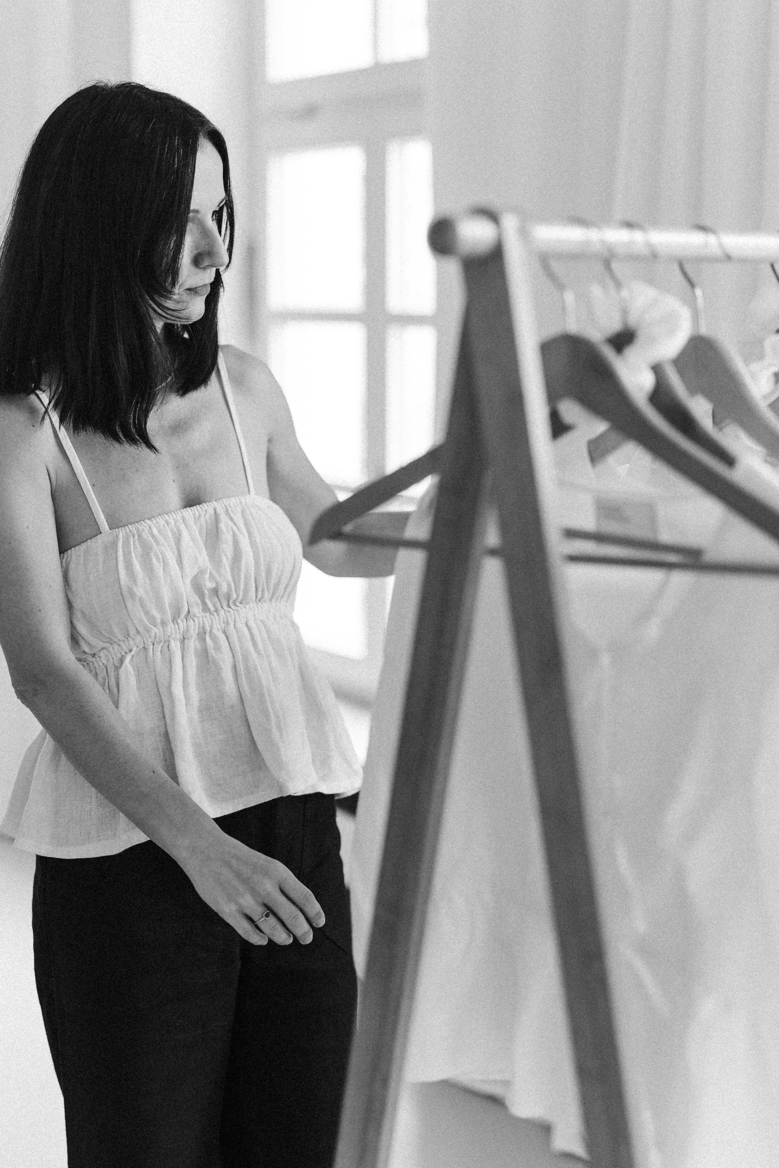 Frau schaut nachdenklich auf eine Kleiderstange mit Hängern in einem Raum mit Fenster im Hintergrund.