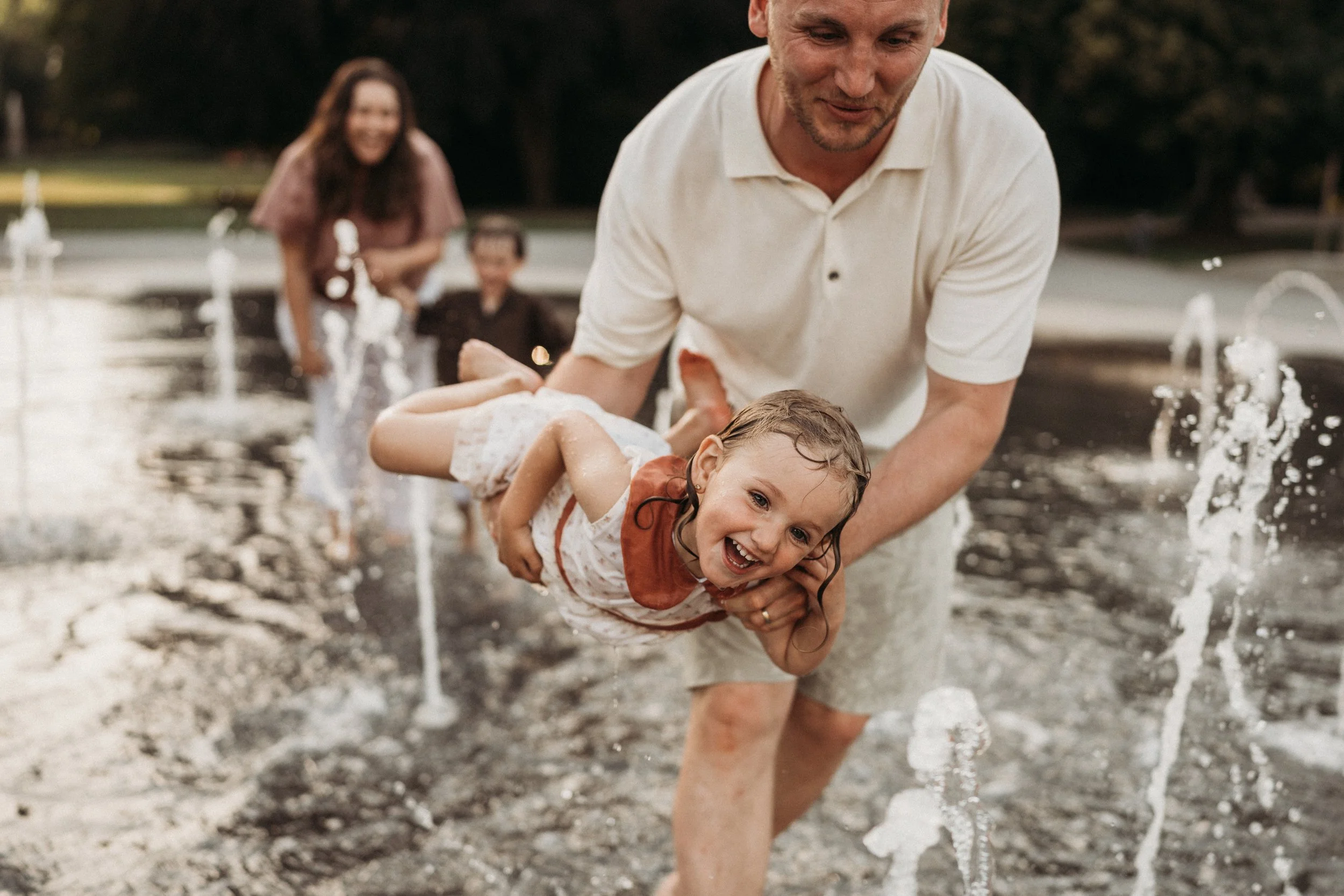Ein Mann hält lachendes Mädchen im Wasser, während im Hintergrund eine Frau und ein weiteres Kind sind.