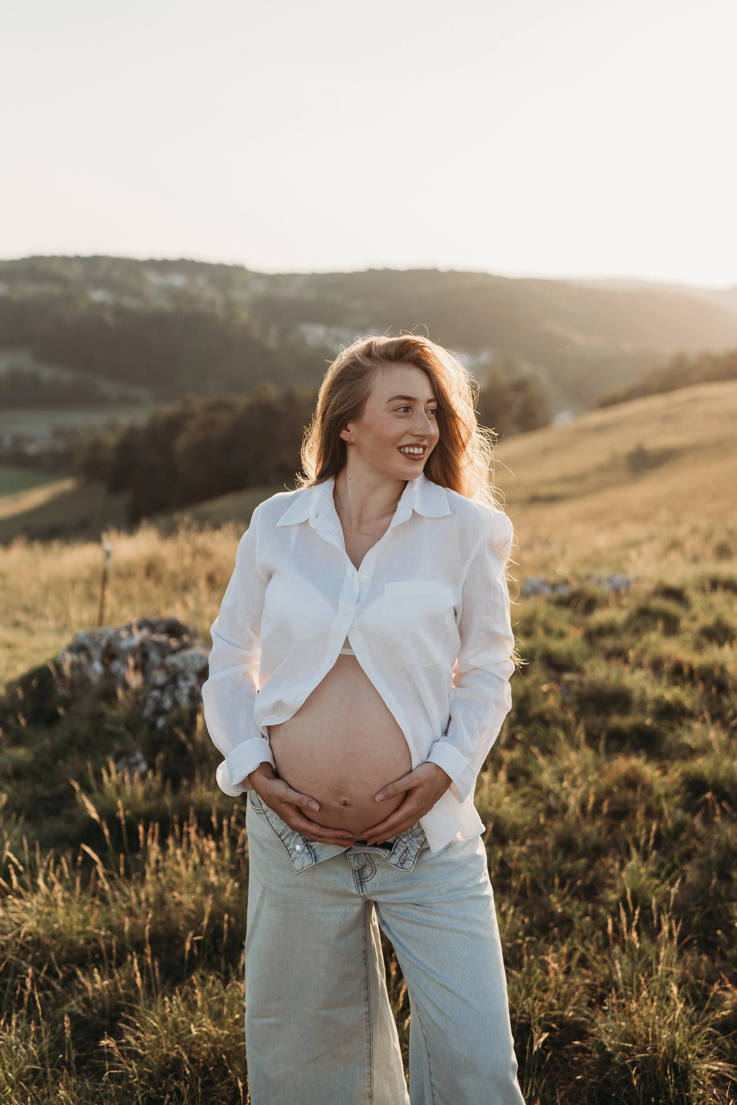 Junge schwangere Frau mit freiem Oberkörper, lockigem Haar, in einer grünen, hügeligen Landschaft bei Sonnenuntergang.