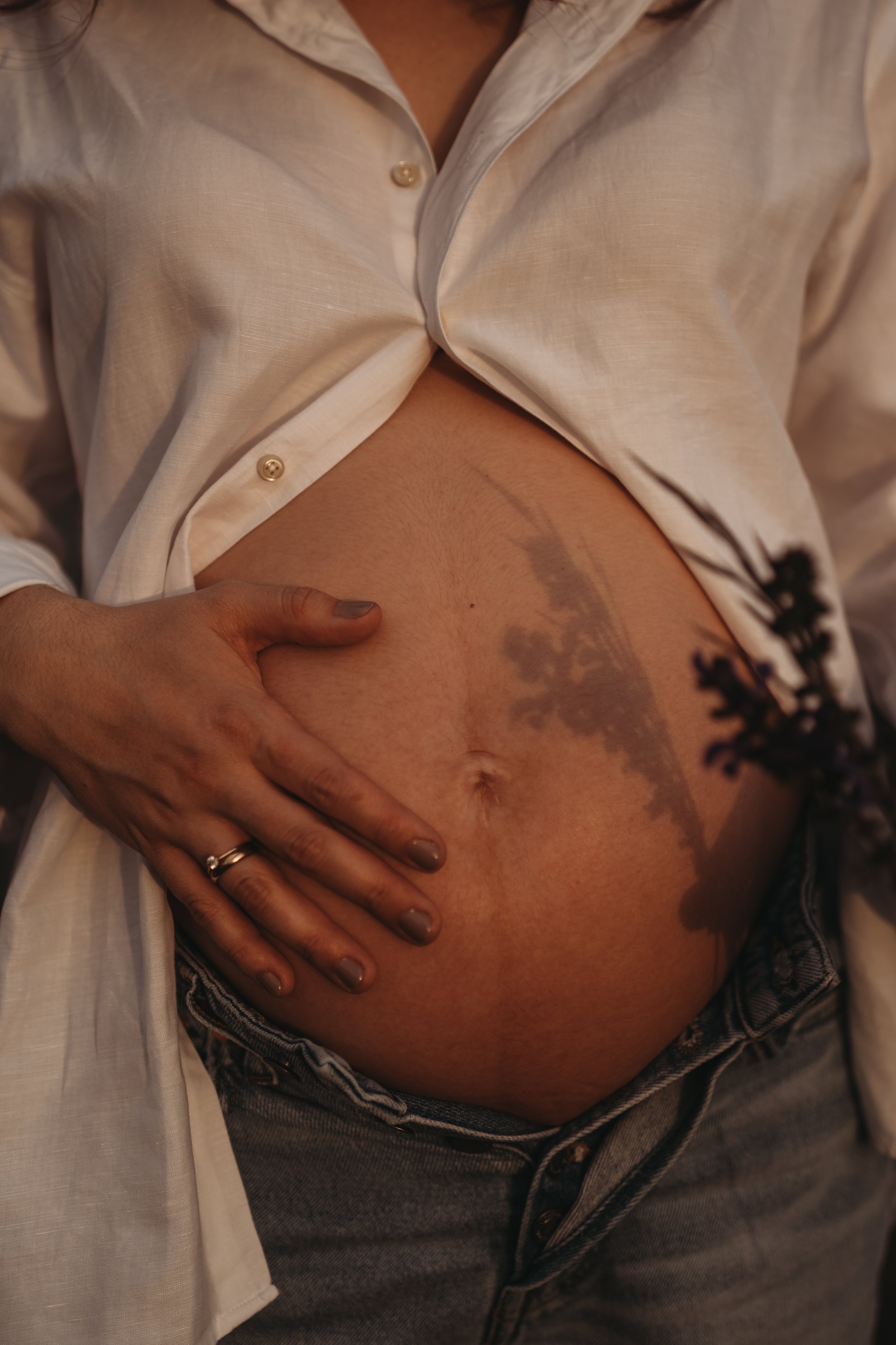 Nahaufnahme eines menschlichen Bauches mit Hand, die den Bauch hält. Das Hemd ist offen, die Haut ist sichtbar, und es gibt eine Tätowierung auf dem Bauch.