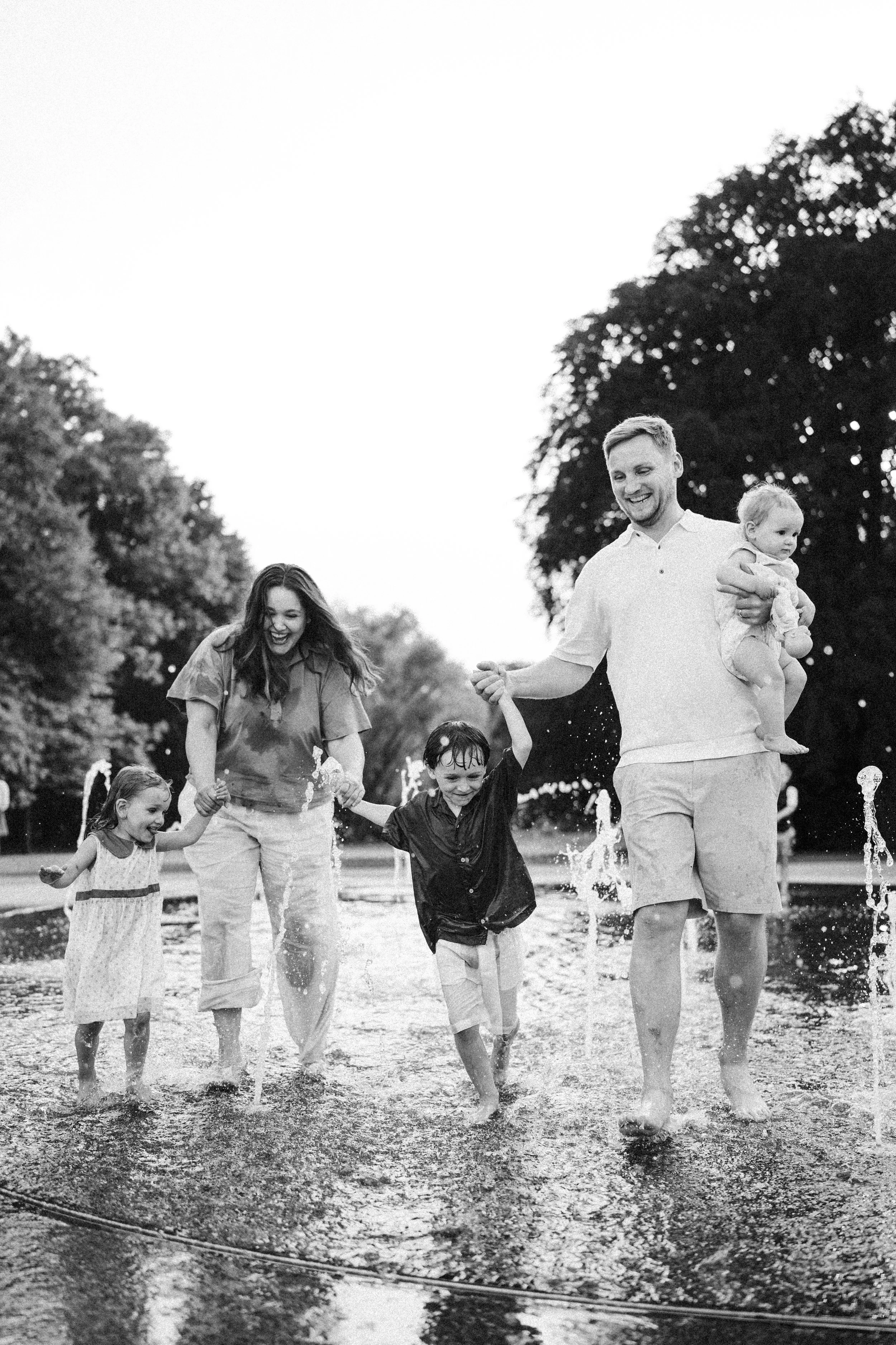 Eine fröhliche Familie läuft barfuß durch eine Pfütze im Freien. Es ist eine Frau, ein Mann und vier Kinder, die lachen und Spaß haben, während sie Wasser spritzen lassen.
