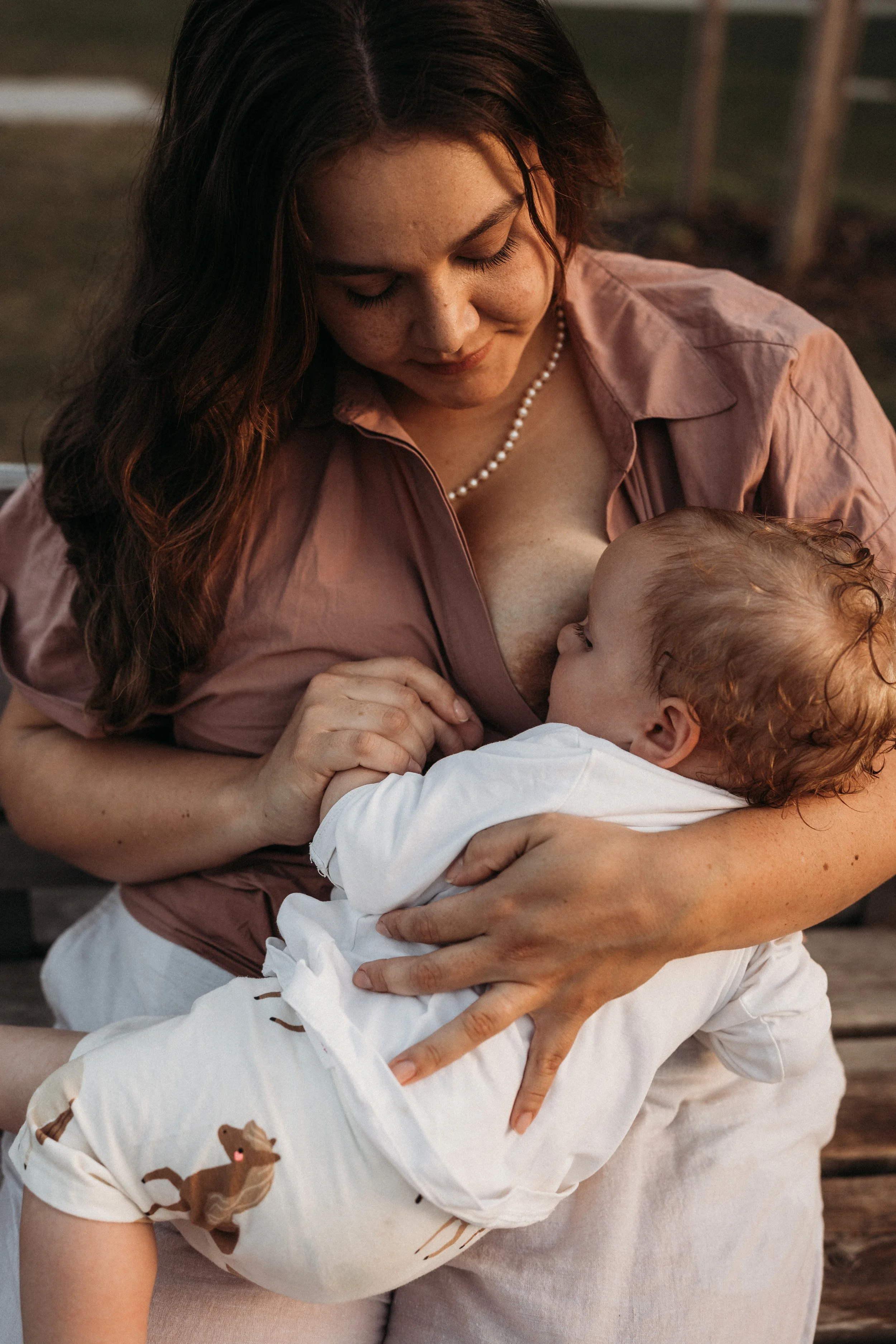 Eine Frau mit braunen Locken und einem Perlenhalsband stillt ein Baby, dessen Gesicht nur teilweise sichtbar ist.