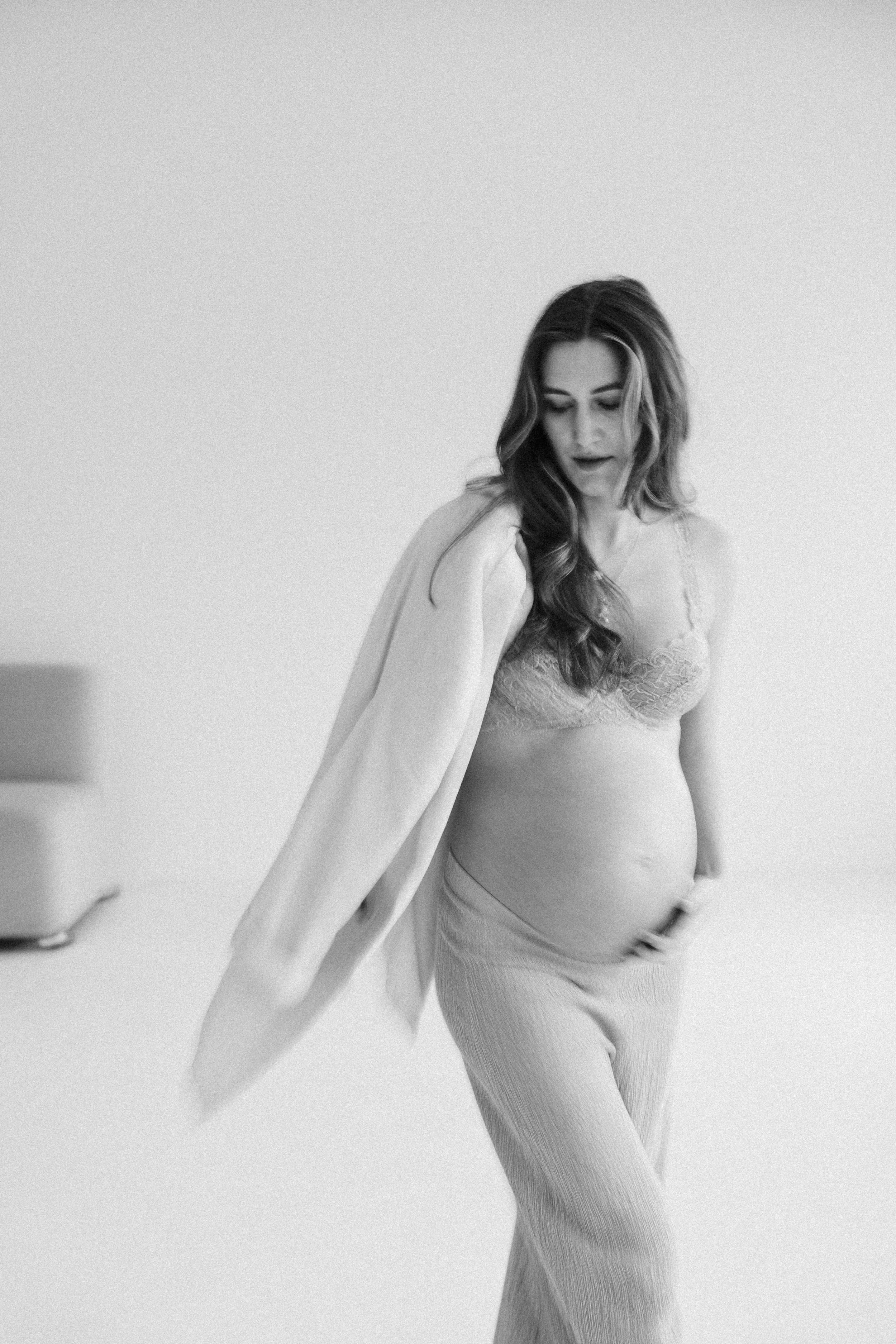 Schwangere Frau in Lingerie und lockerer Hose, posiert in einem minimalistischen Raum, schwarz-weiß Fotografie.