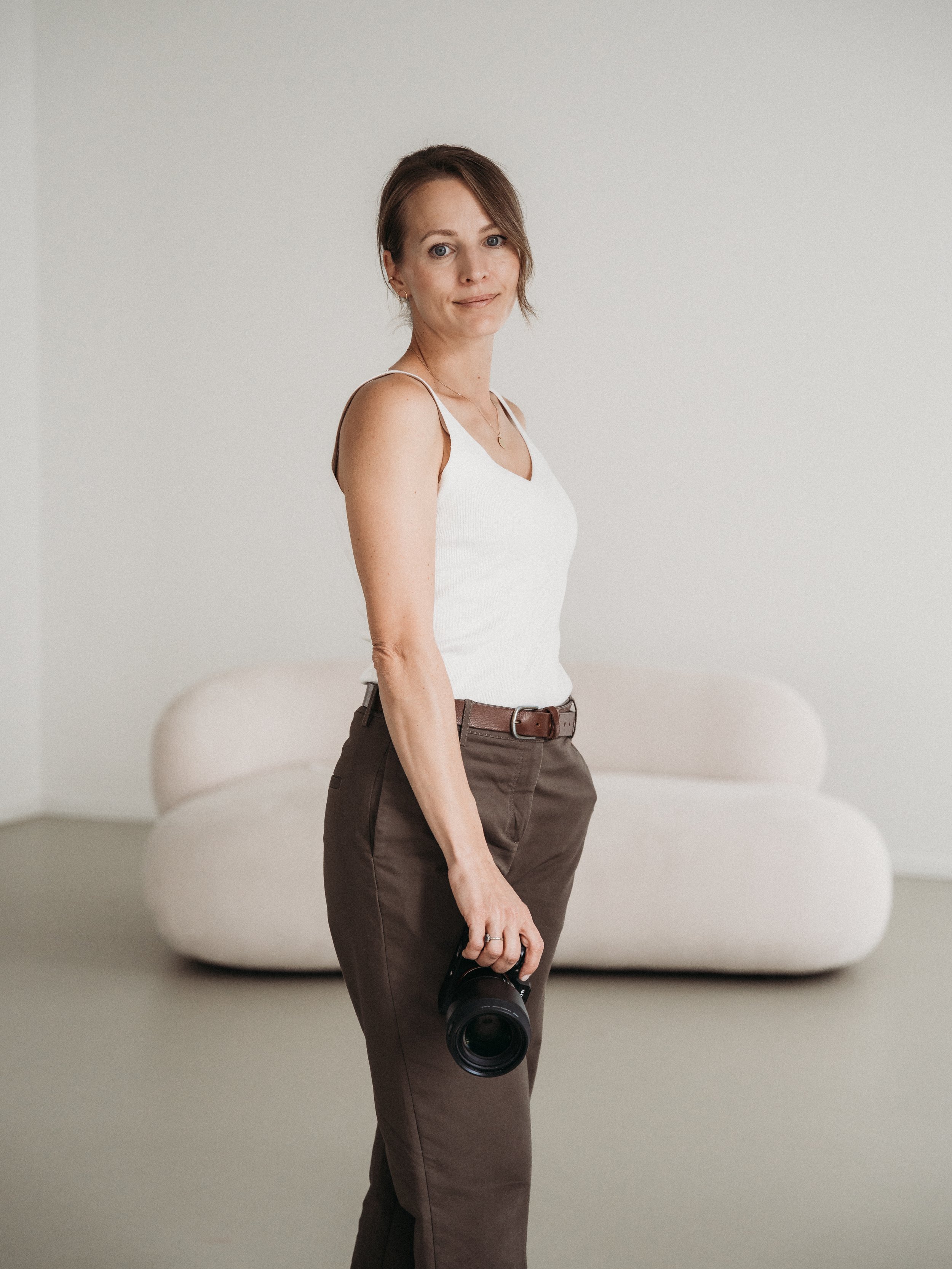 Eine Frau steht vor einem weißen Sofa, trägt ein weißes Top, braune Hose, Gürtel und hält eine Kamera. Sie hat kurze braune Haare und schaut in die Kamera.