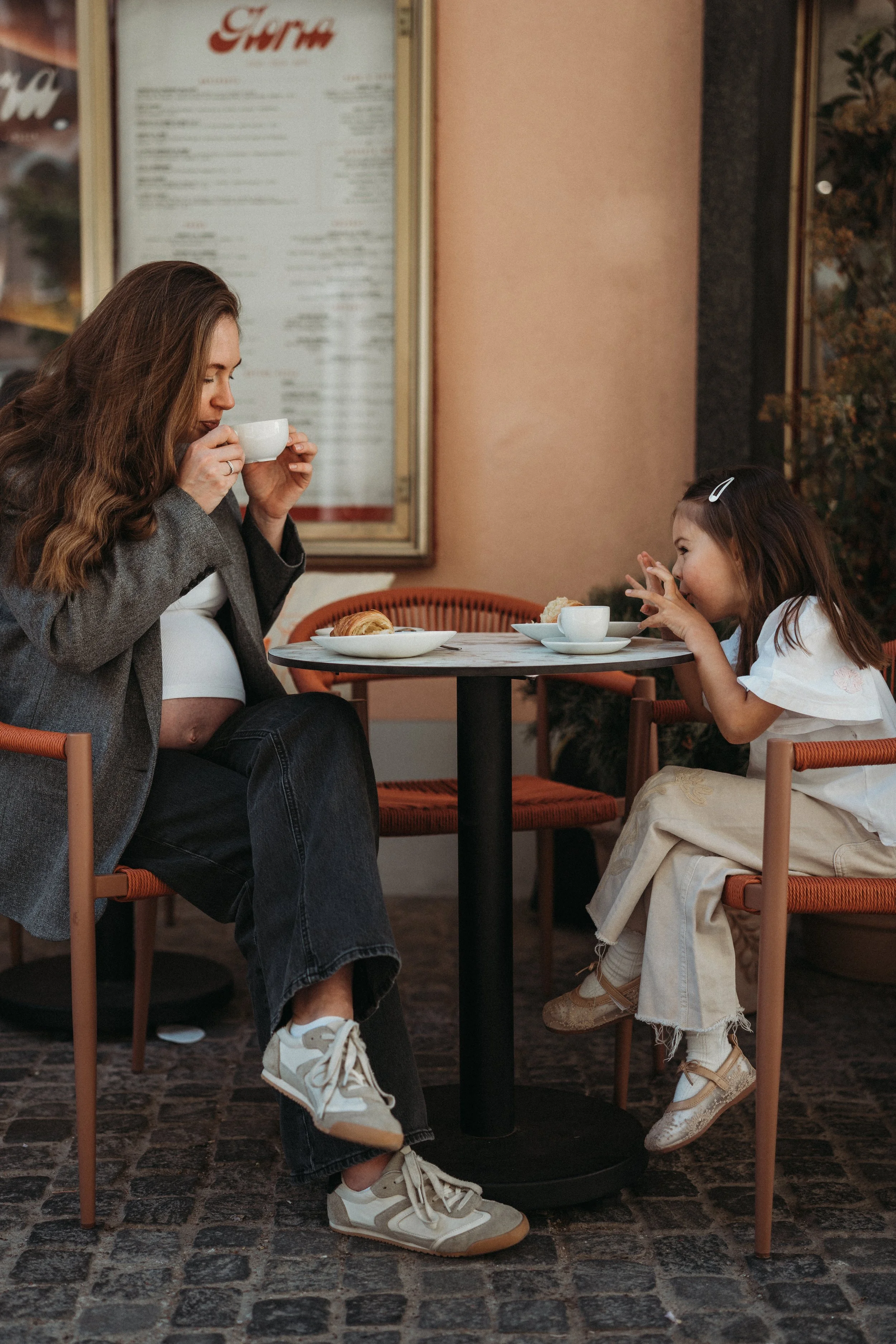 Eine Frau und ein Mädchen trinken Kaffee in einem Café im Freien, während sie auf einem runden Tisch sitzen. Das Mädchen trägt einen weißen Rock und Schuhe, die Frau trägt einen grauen Blazer und Sneakers.