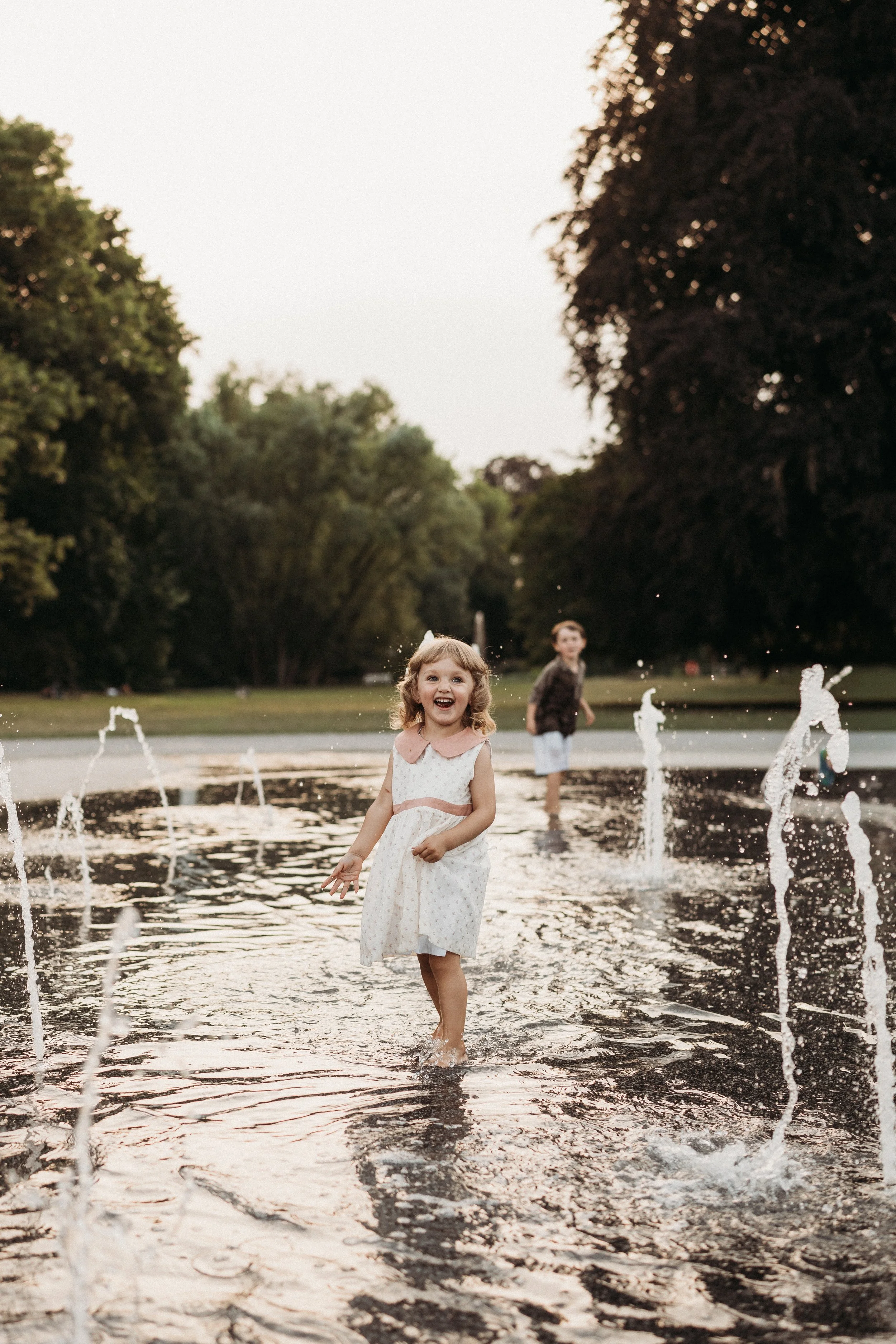 Ein lachendes Mädchen läuft barfuß durch einen Wasserbrunnen in einem Park, während im Hintergrund ein Junge steht. Es ist Abend und die Bäume umgeben die Szenerie.