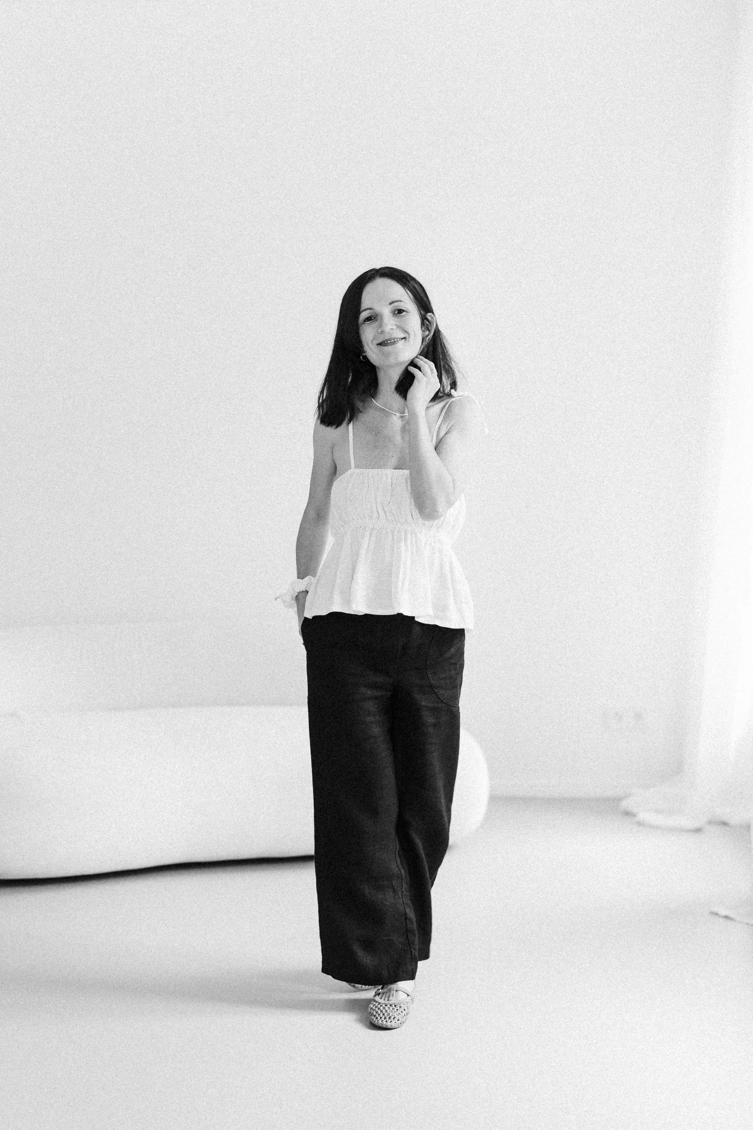 Schwarz-weiß-Fotografie einer Frau, die lächelnd in einem hellen Raum steht, trägt eine weiße Top und schwarze Hosen, mit einer Hand am Kinn.