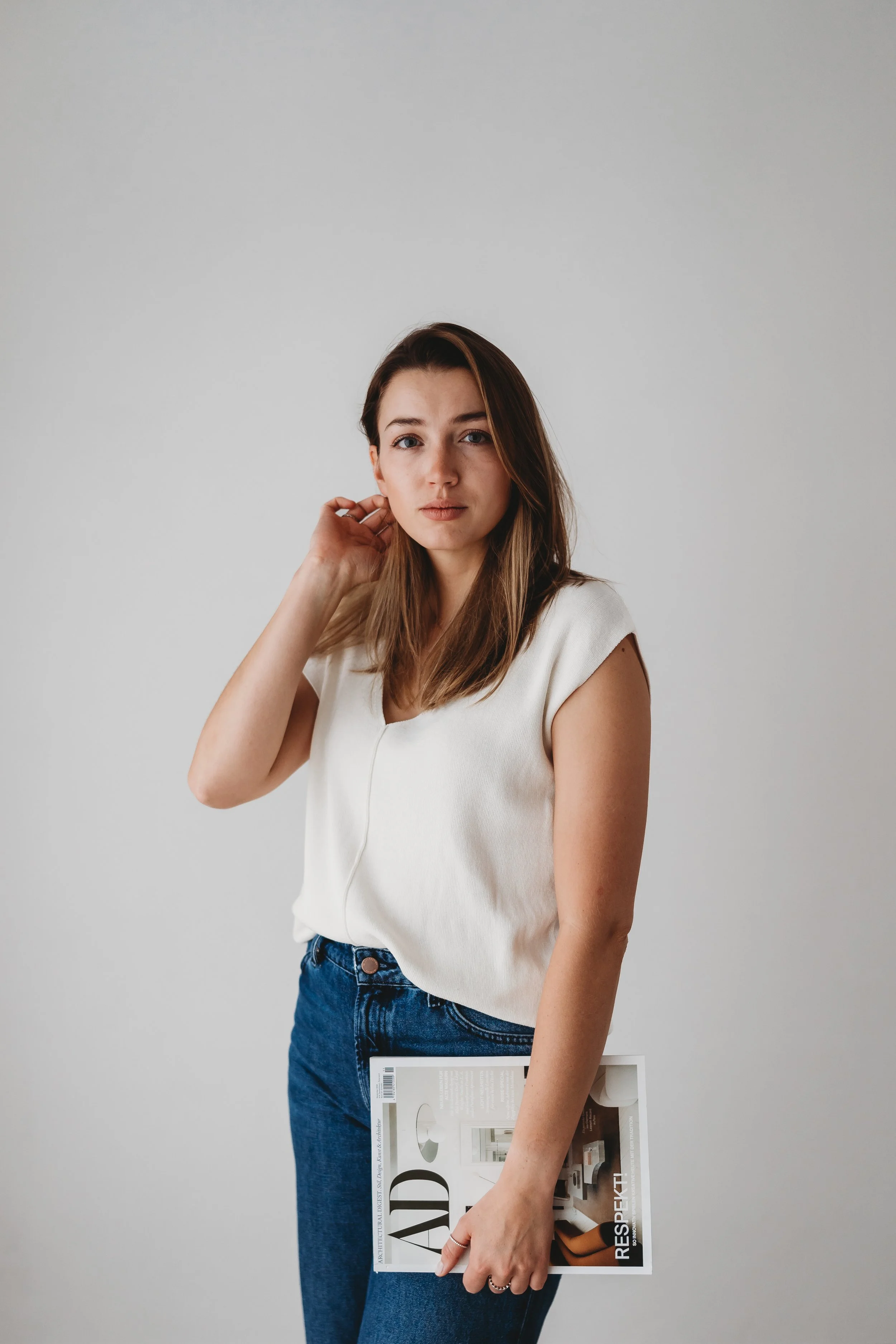 Junge Frau mit braunen Haaren trägt ein weißes Oberteil und blaue Jeans, hält eine Zeitschrift und steht vor einem neutralen grauen Hintergrund.
