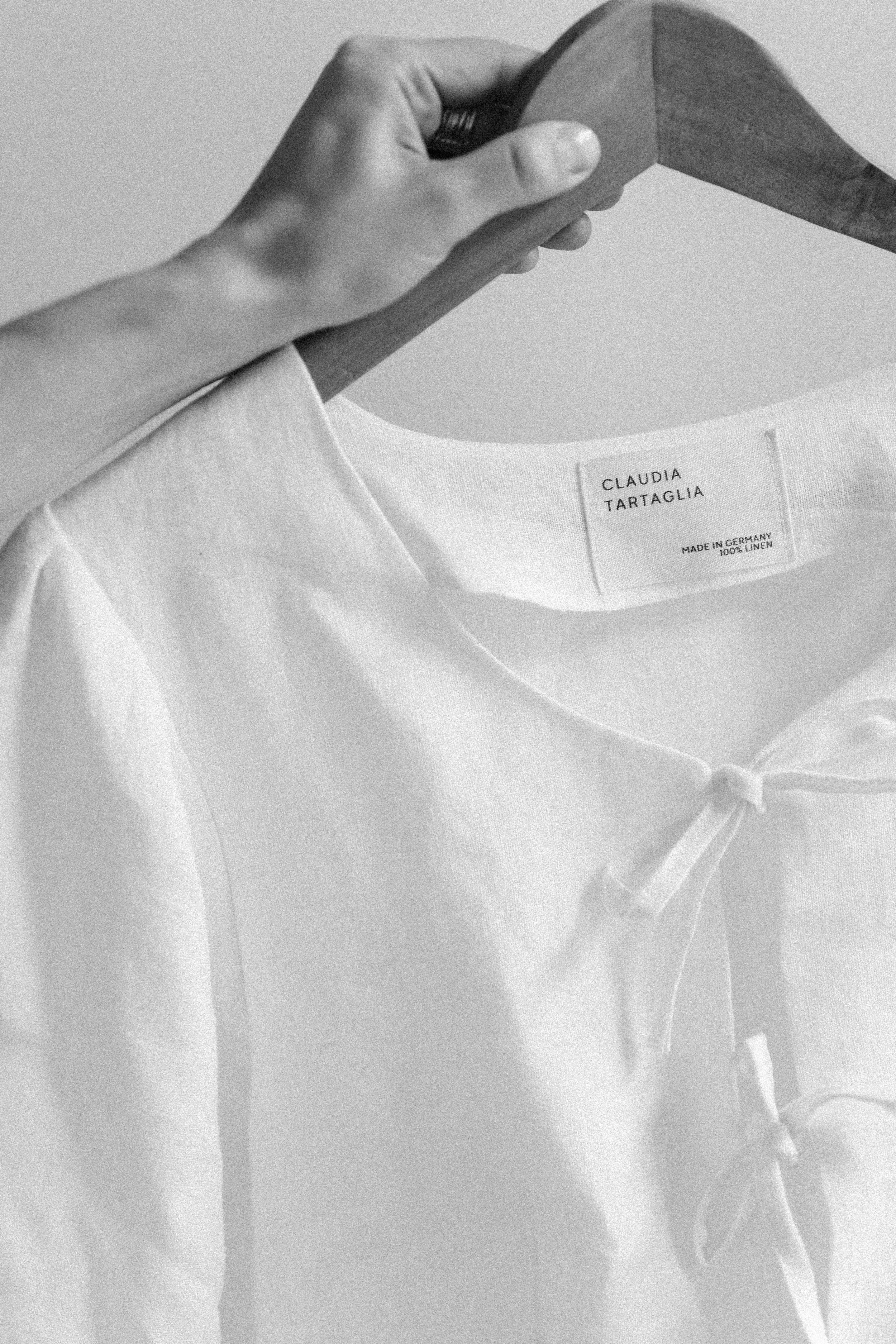 Ein weißes Hemd mit einem kleinen Knoten an der Kragenöffnung an einer Holz-Kleiderstange. Der Hemd-Etikett zeigt den Namen 'Claudia Tartaglia' und die Angaben 'Made in Germany, 100% Linen'.