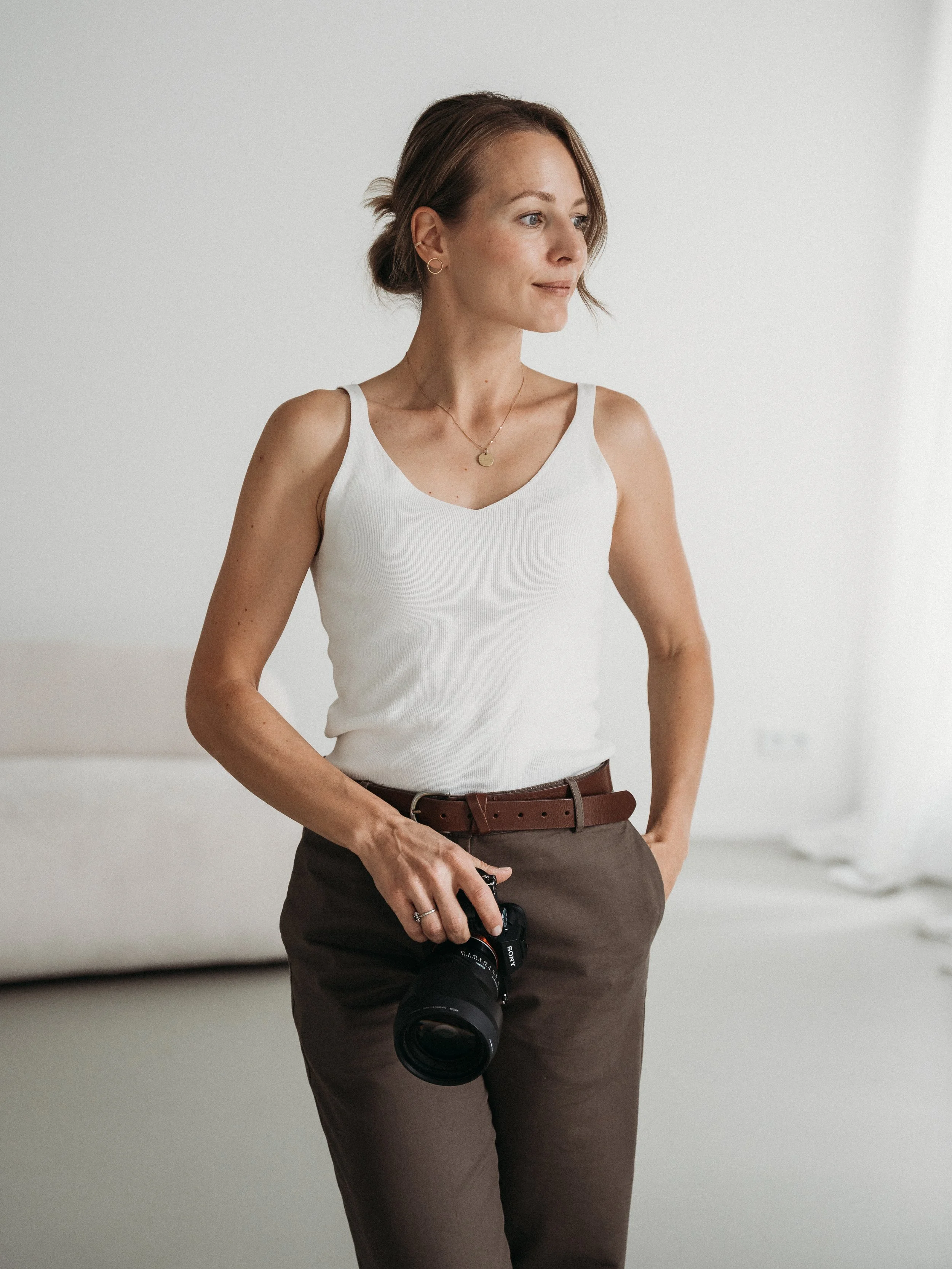 Eine Frau trägt weiße ärmellose Bluse, braune Hose, braunen Gürtel und trägt eine Kamera. Sie hat kurze braune Haare und trägt kleine Goldohrringe und eine Halskette.