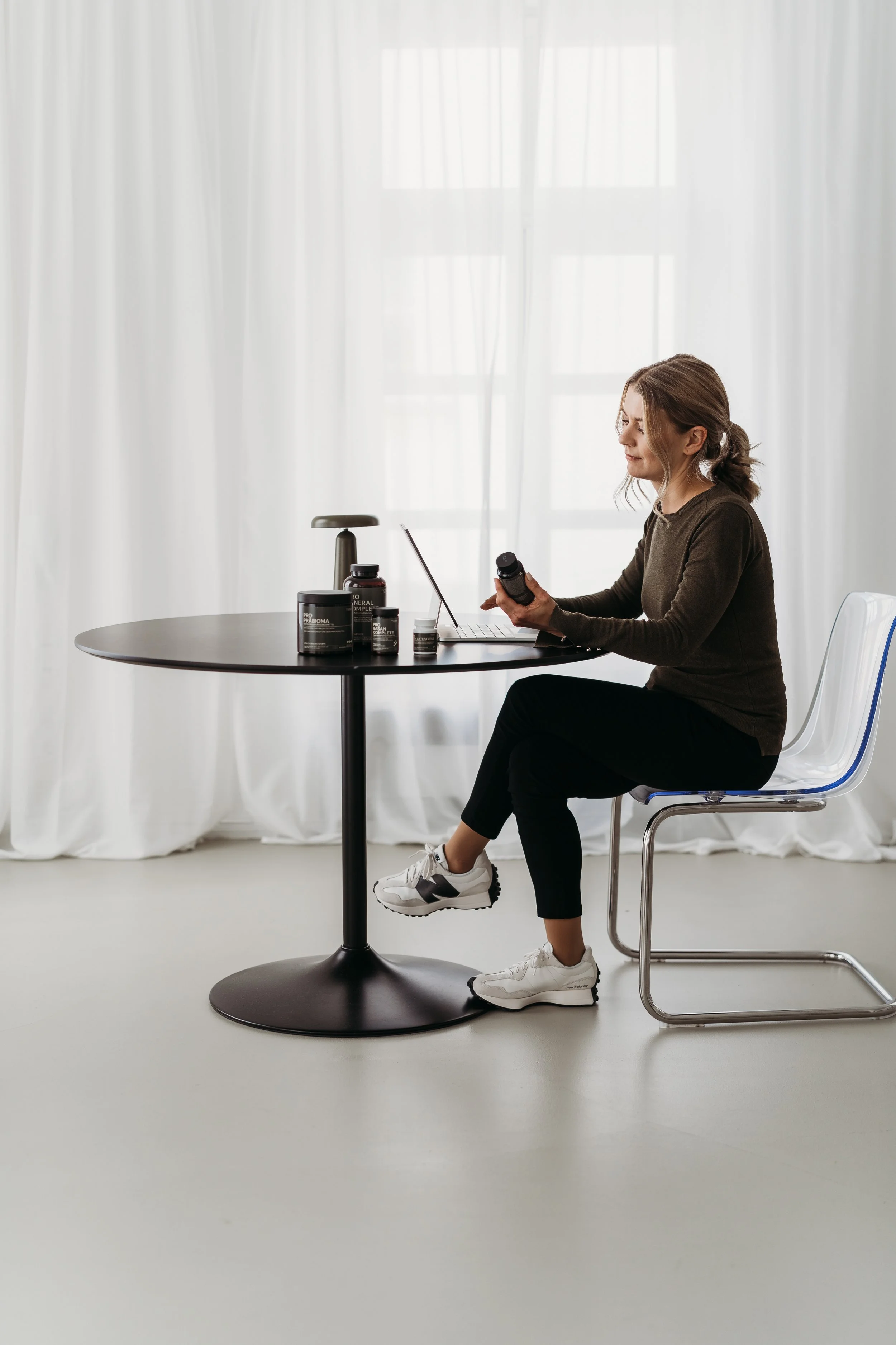 Frau sitzt an einem Tisch mit Nahrungsergänzungsmitteln, hält eine Flasche und schaut auf einen Laptop, im Hintergrund weiße Vorhänge.