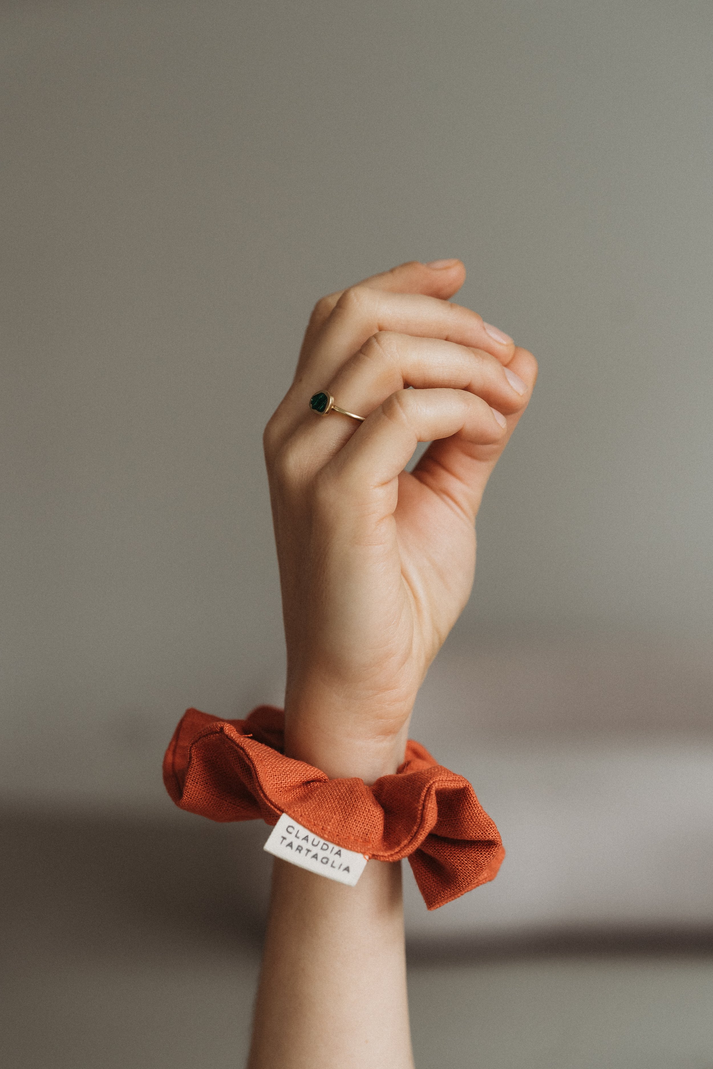 Eine Hand mit einem Goldring, getragen an einem Ringfinger, und einem orangefarbenen Stoffarmband mit einem weißen Etikett, auf dem steht 'ClaudiA Tartaglia'.