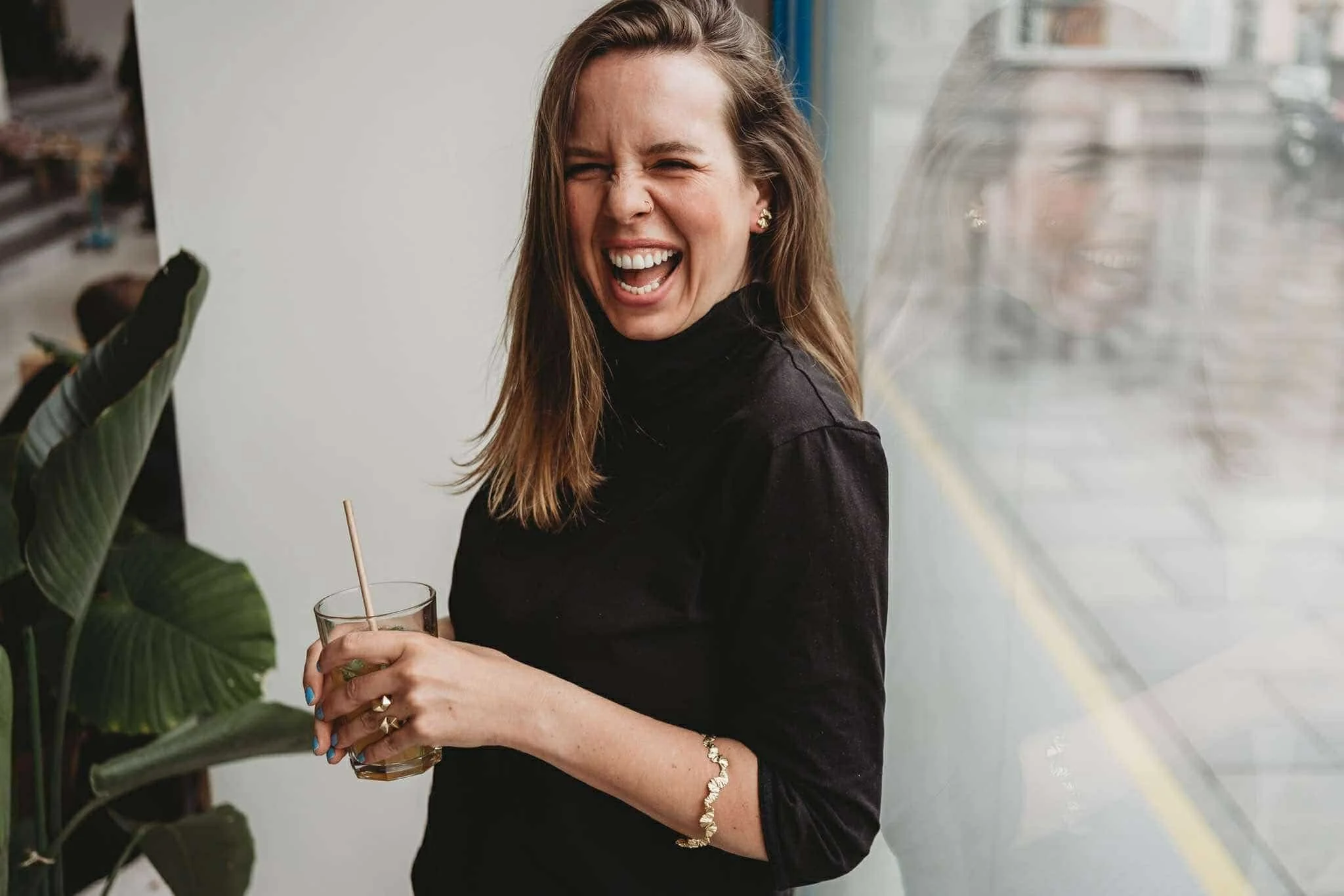 Frau in schwarzem Pullover lacht und hält ein Getränk in der Hand. Sie steht neben einer großen Pflanze und einem Fenster, in dem ihr Spiegelbild sichtbar ist.