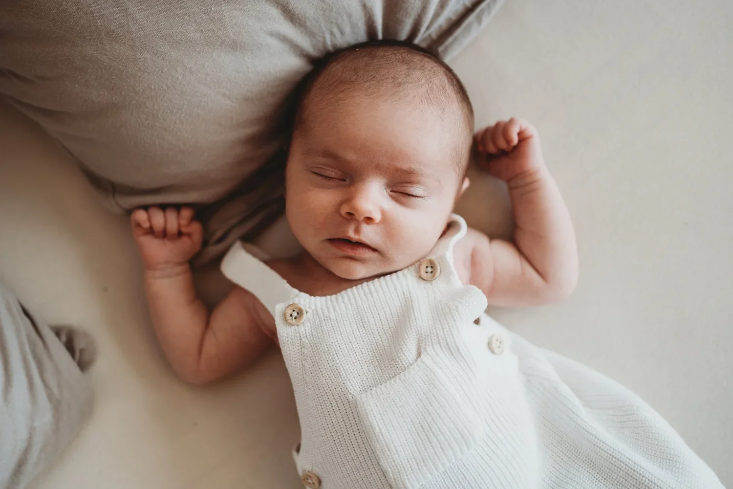 Ein schlafender Säugling liegt auf einem weichen, hellen Kissen. Das Baby trägt eine weiße Strickweste mit Knöpfen und hat kurze, dunkle Haare.