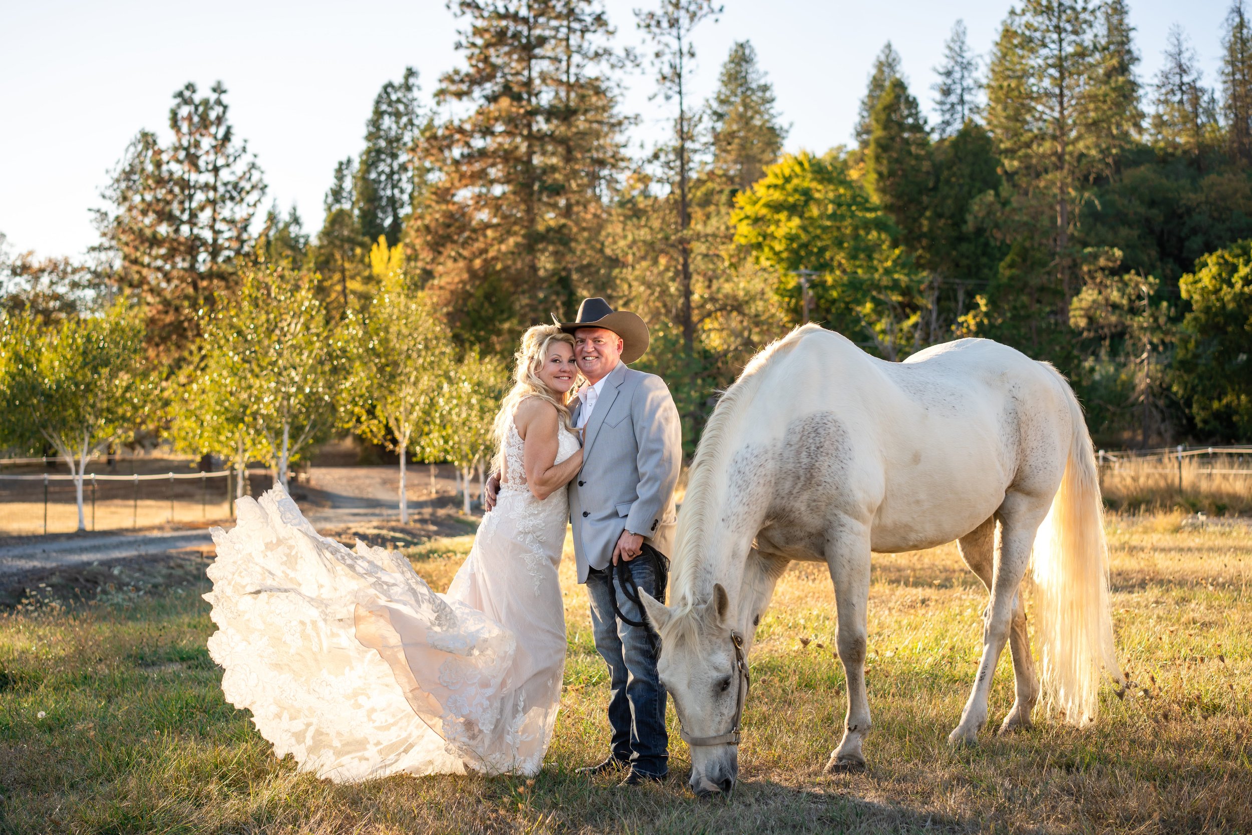 Outdoor wedding reception area with rustic wooden buildings, a welcome sign, a high-top table with decor, and a scenic landscape with trees and sunlight.