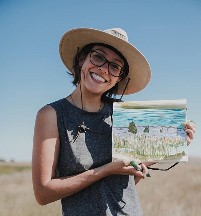 Young woman smiling, wearing a wide-brimmed hat and glasses, holding a sketchbook with a landscape drawing outdoors on a sunny day