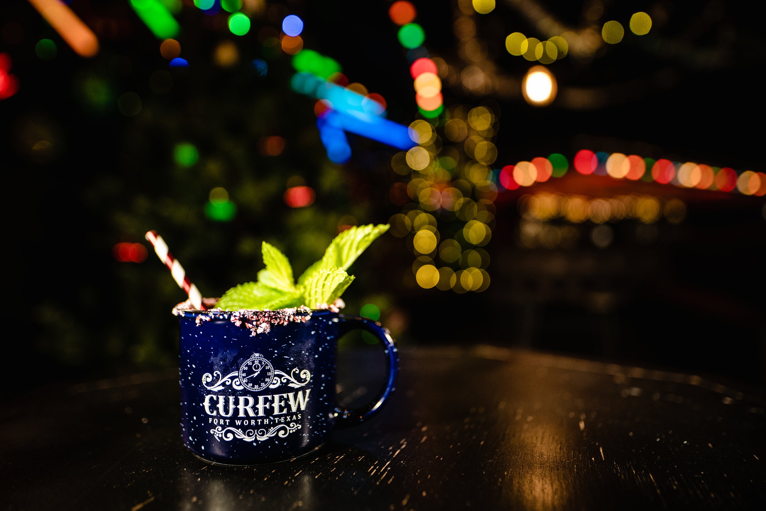 Blue mug with "CURFEW Fort Worth, Texas" logo, garnished with mint leaves and a candy cane straw, on a wooden table with colorful bokeh lights in the background.