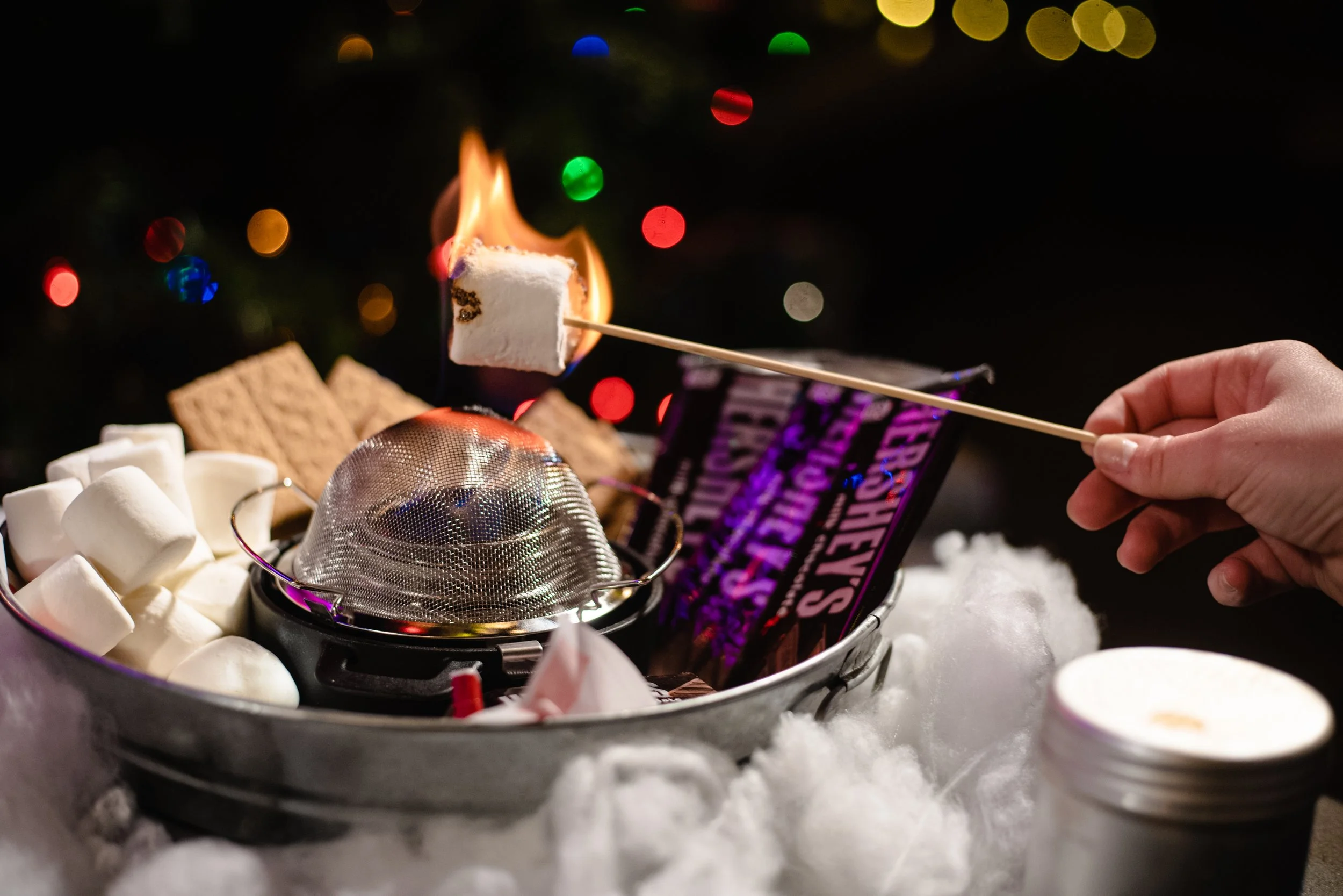 Person roasting a marshmallow over a flame with s'mores ingredients, including graham crackers and chocolate, set in a holiday-themed setting with colorful lights.