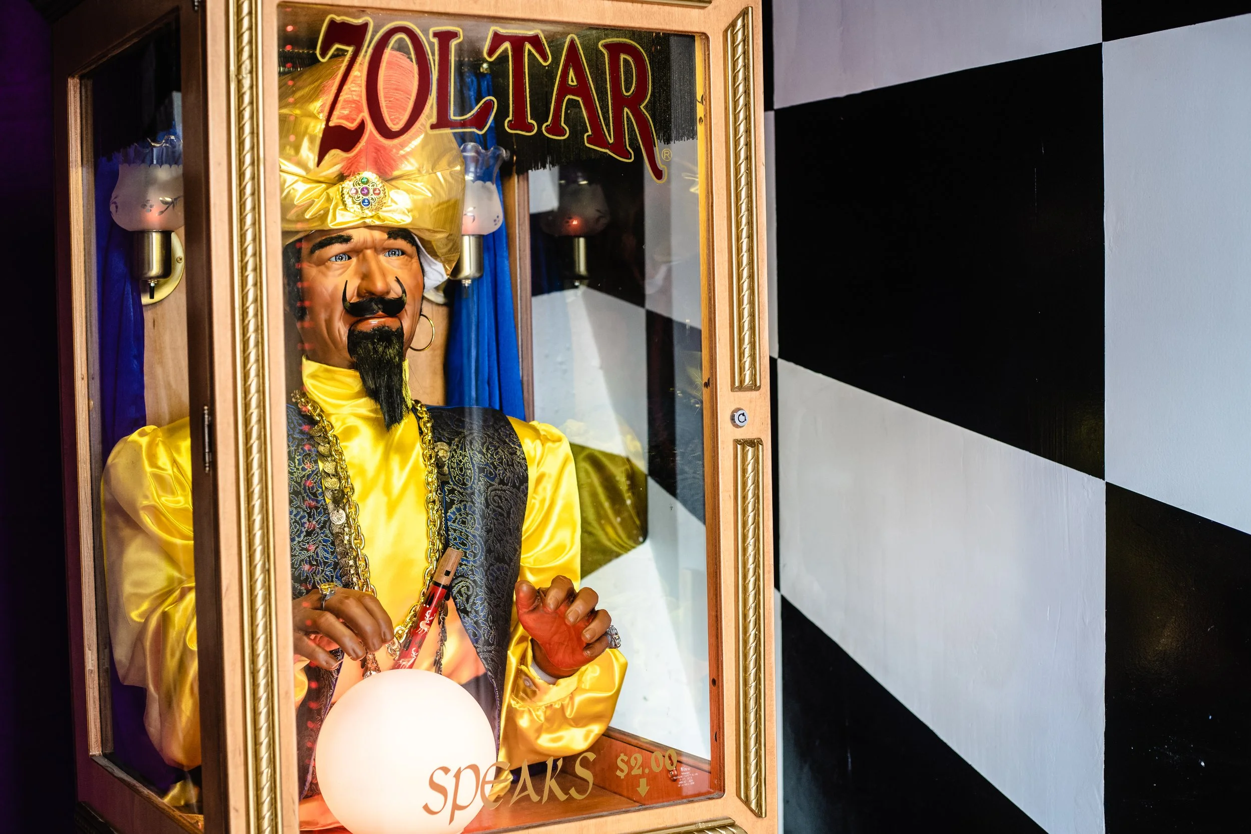 Zoltar fortune-telling machine with a mannequin inside, displaying a glowing crystal ball, in front of a black and white checkered wall.