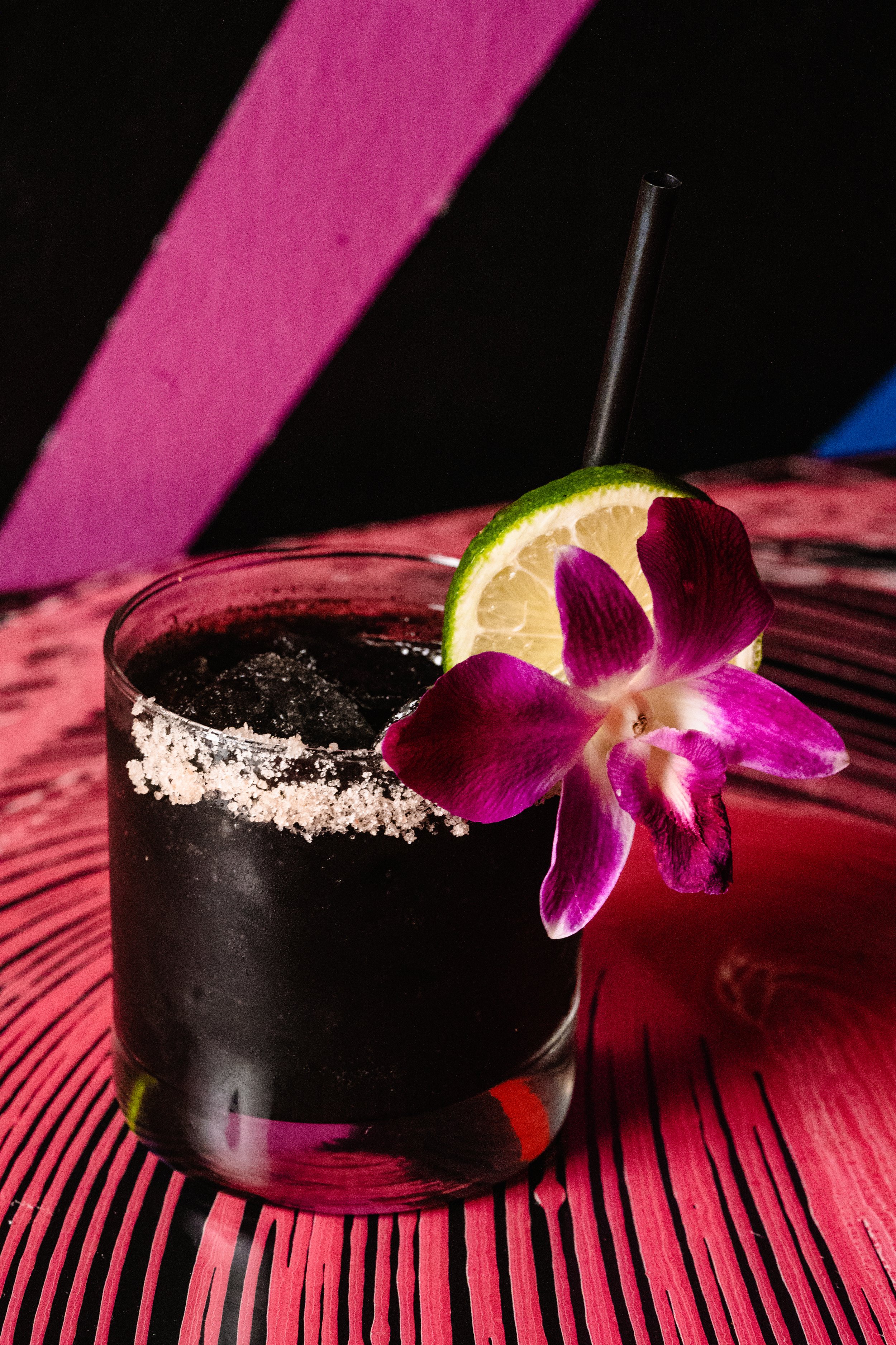 Black cocktail in a glass with a salted rim, garnished with a lime slice and purple orchid on a colorful striped table.