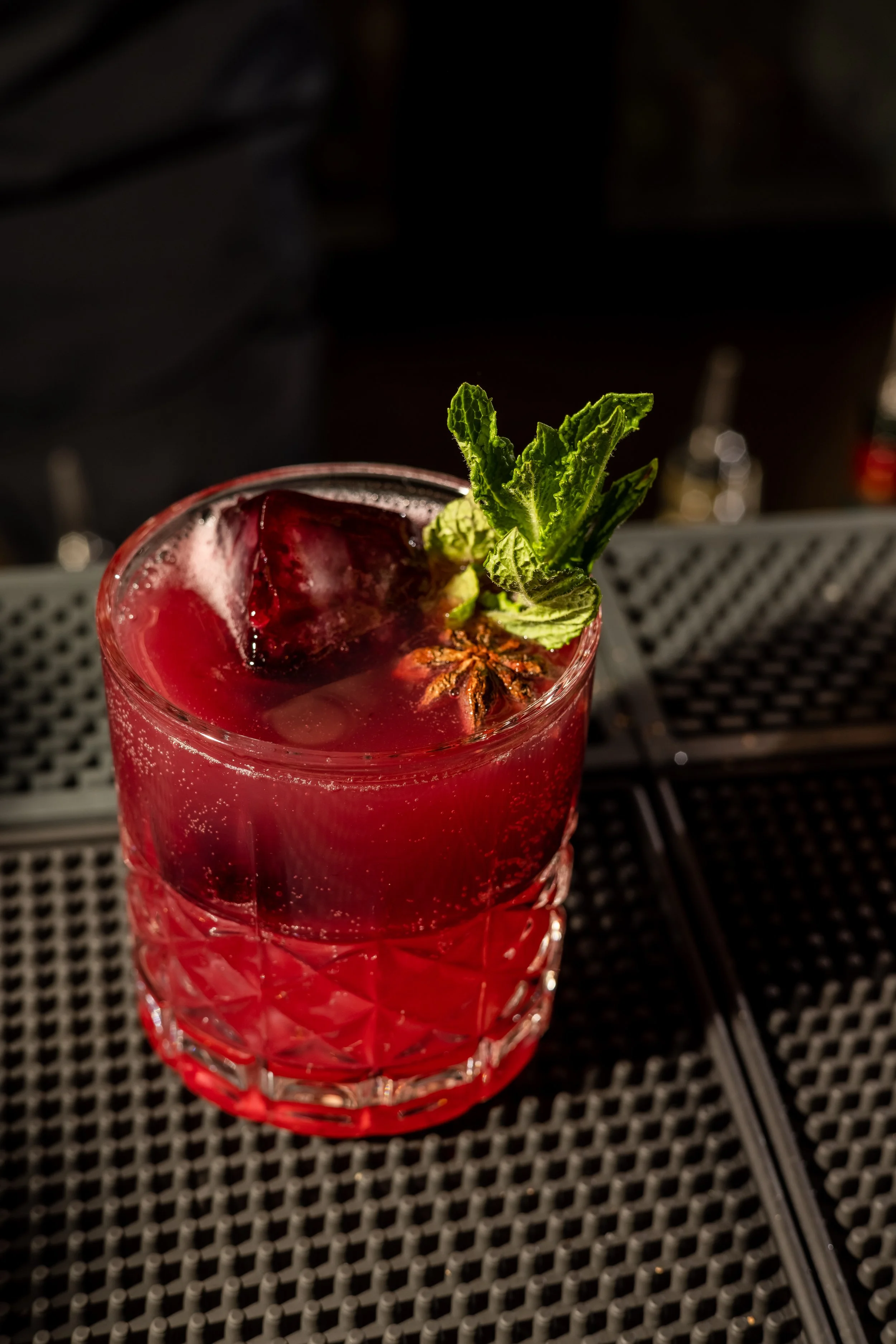 A red cocktail in a glass with ice, garnished with fresh mint leaves and a star anise, on a bar mat.