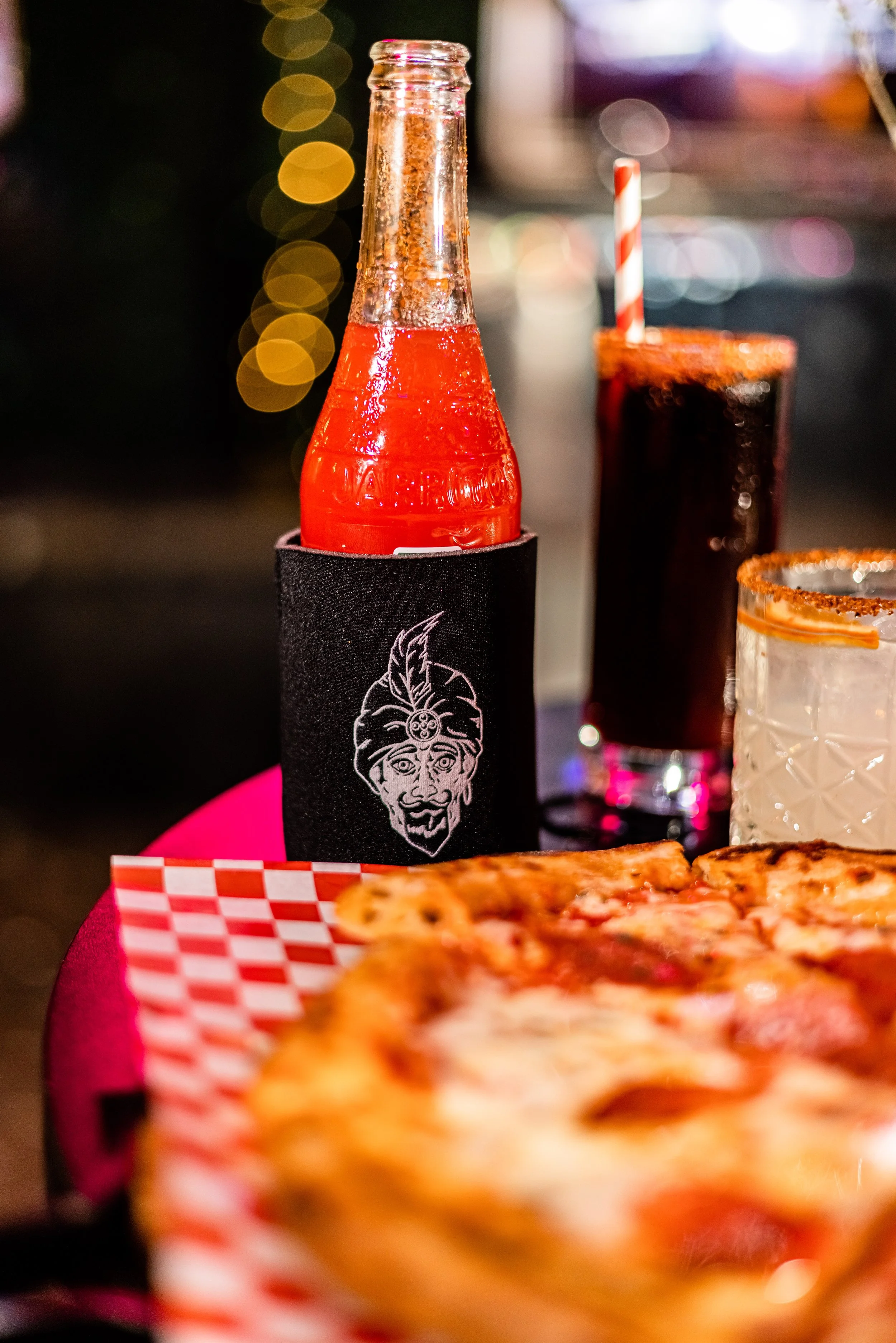 A scene featuring a bottle of orange soda wrapped in a black sleeve with an illustration, a glass of dark soda with a red and white straw, a cocktail with a salt rim, and a pizza on a red checkered paper.