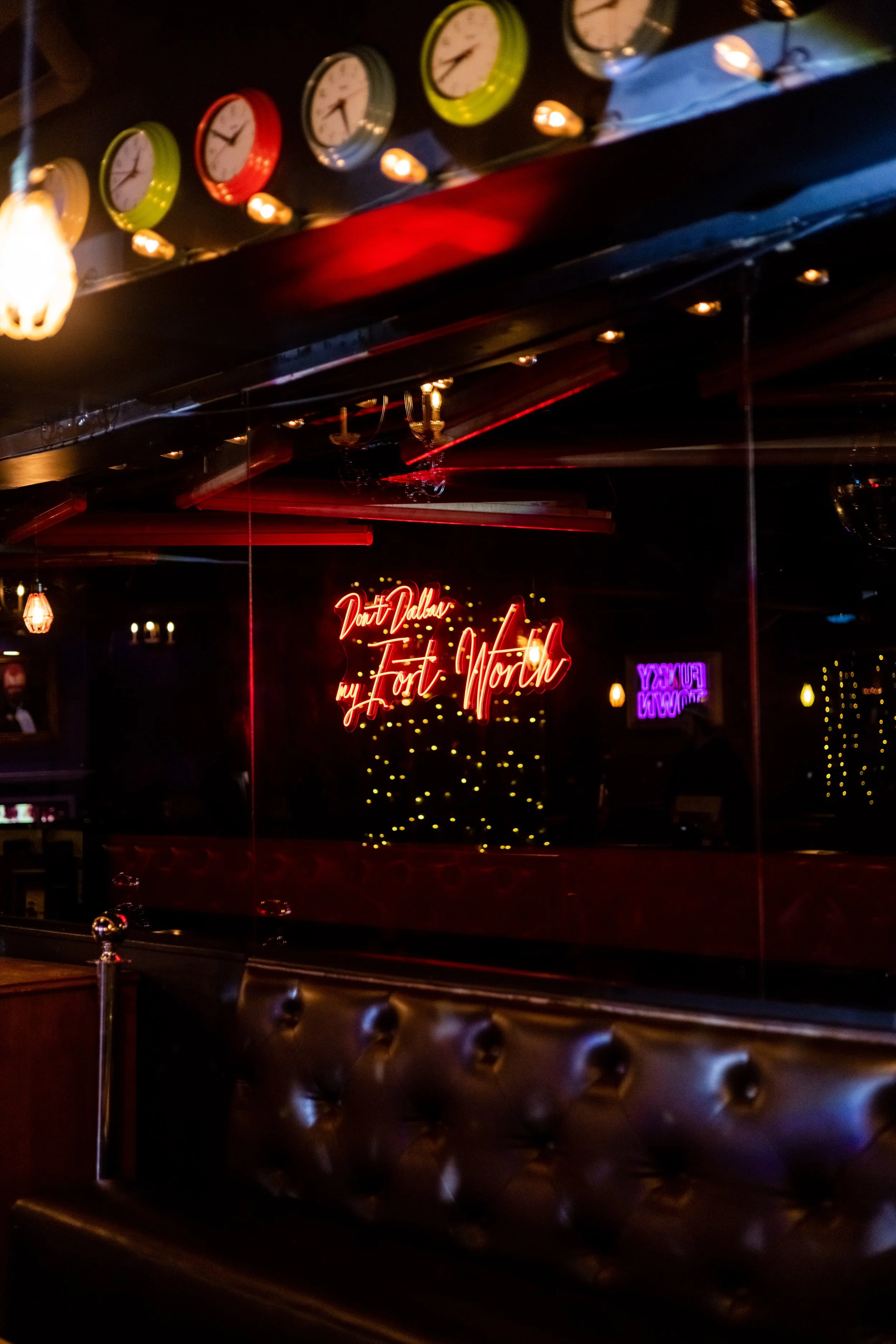 Dimly lit bar interior featuring a tufted black leather booth, colorful clocks on the wall, and a neon sign reading "Don't Dallas my Fort Worth."