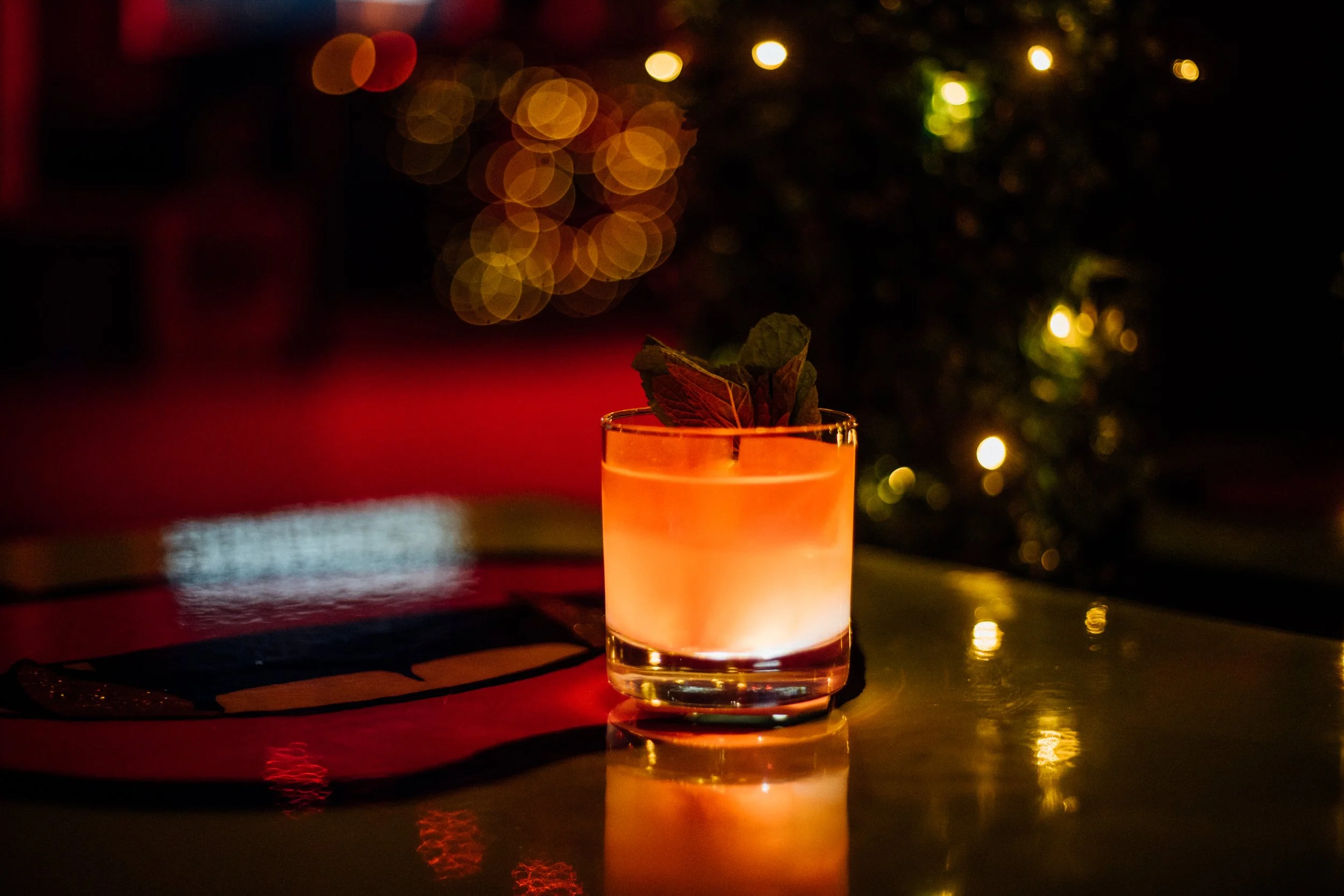 A cocktail with mint garnish on a table, surrounded by blurred lights in a dimly lit bar.