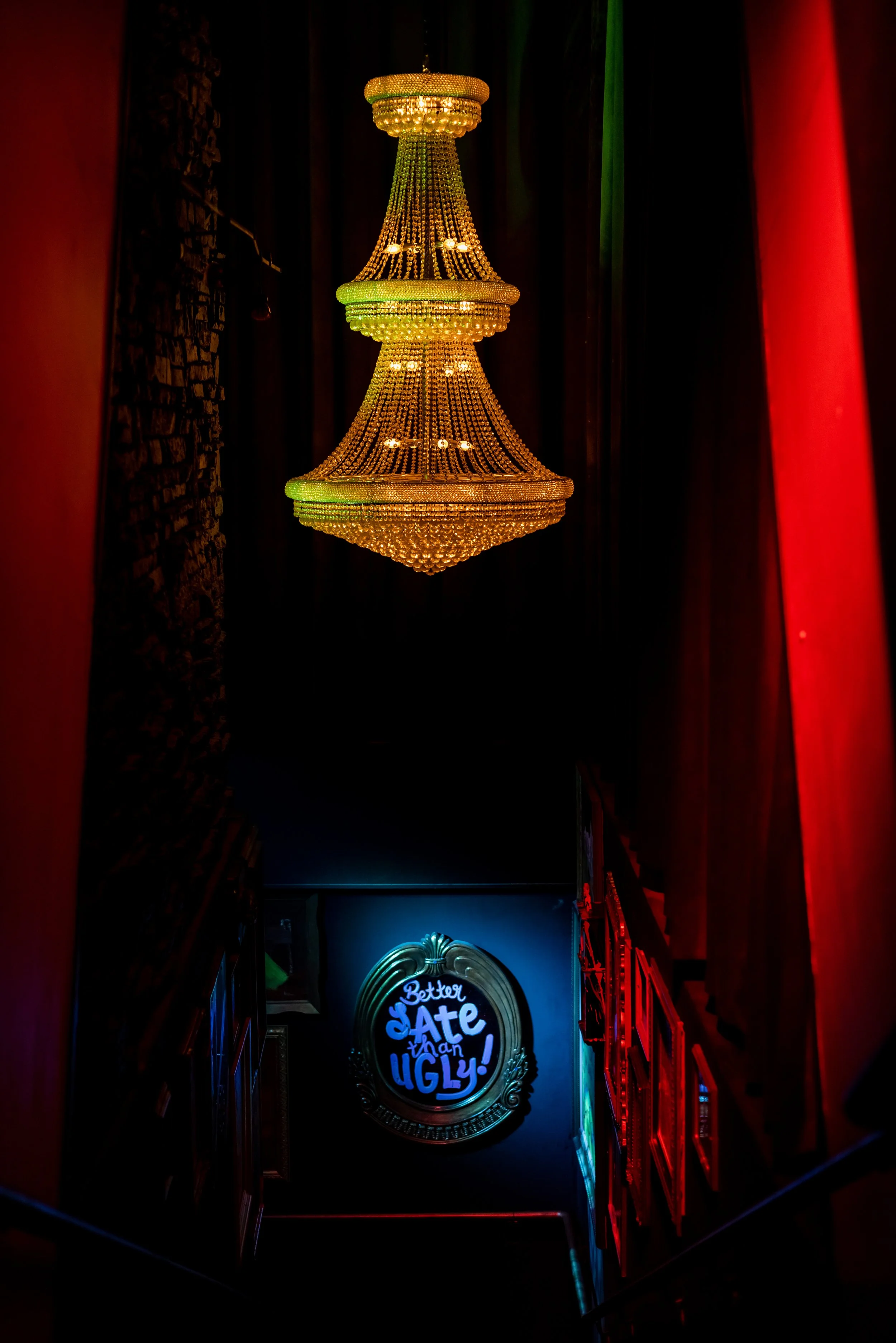 Dimly lit stairway with a large, ornate chandelier and a neon sign that says 'Better Late Than Ugly.'