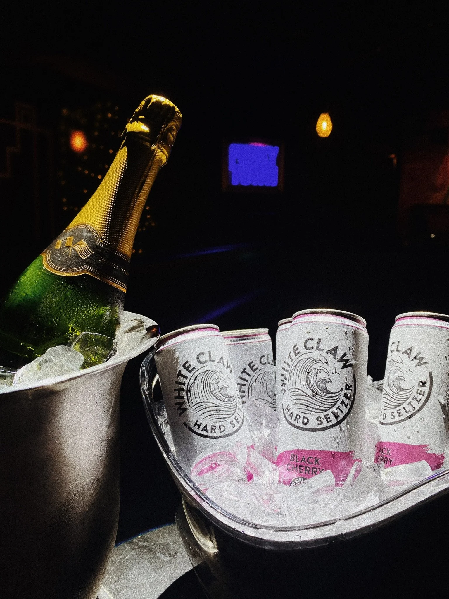 A bottle of champagne in an ice bucket next to several cans of White Claw Black Cherry hard seltzer in another ice-filled container, on a dimly lit table.