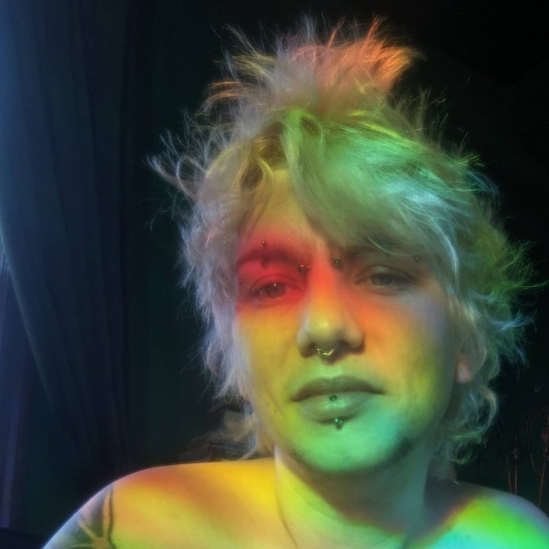 selfie of rose, a white non-binary person with messy blonde hair and a lot of piercings. they're in front of a dark green curtain and red and green light from a prism shines on their face.