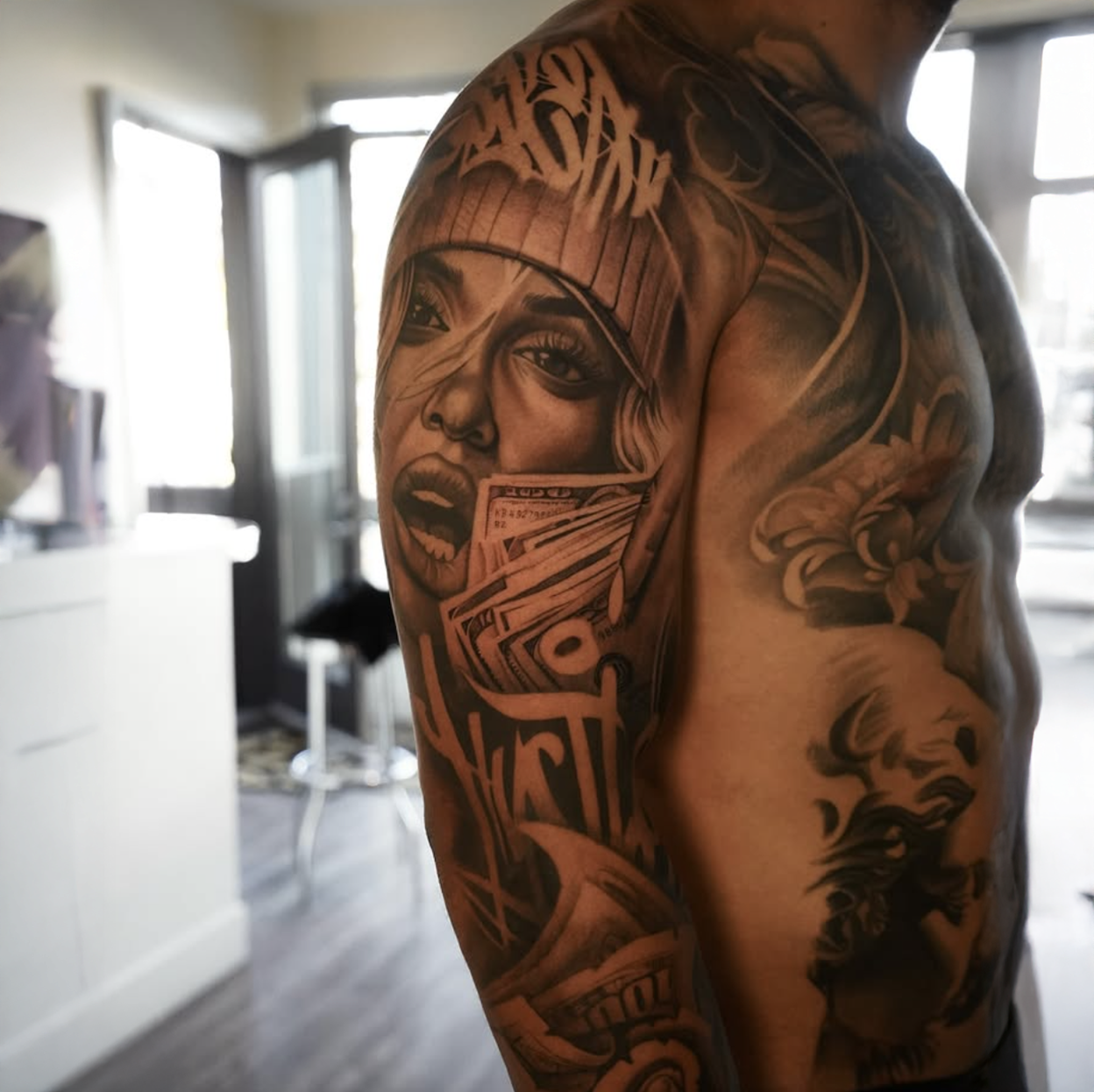 Tattoo of a woman's face with flowing hair, holding dollar bills, on a man's arm and shoulder, with a tattooed sleeve on his upper arm.