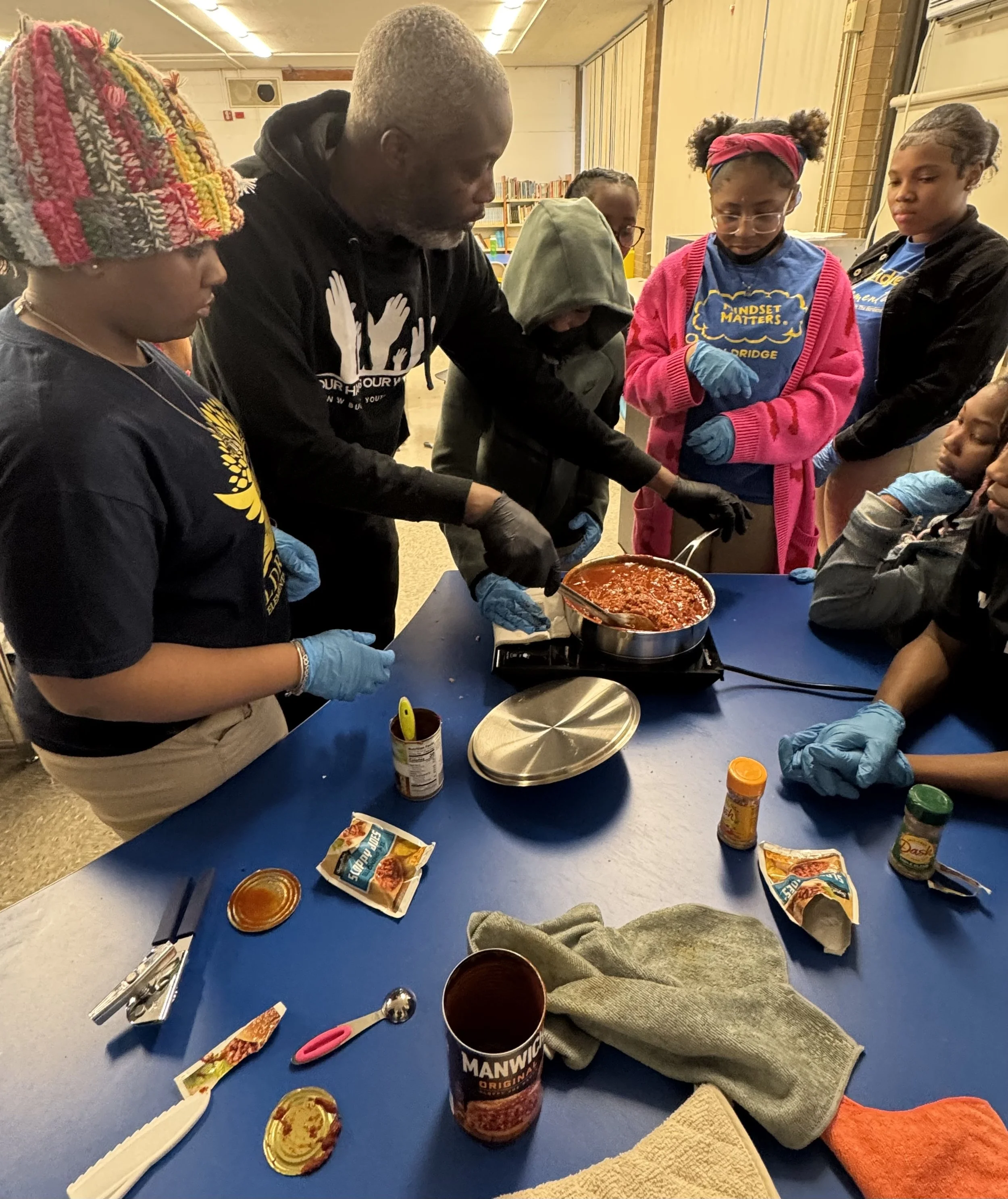 Group of children and an adult gathered around a table with a pan of chili, involved in a cooking activity. The table has utensils, spice packets, and cans on it.