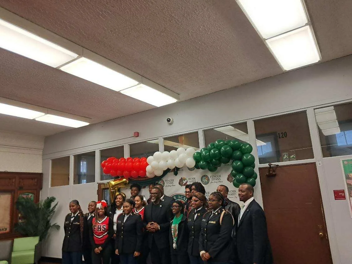 Group of people posing for a photo indoors, with balloons and a banner decorated in red, white, and green in the background, suggesting a celebration or event.