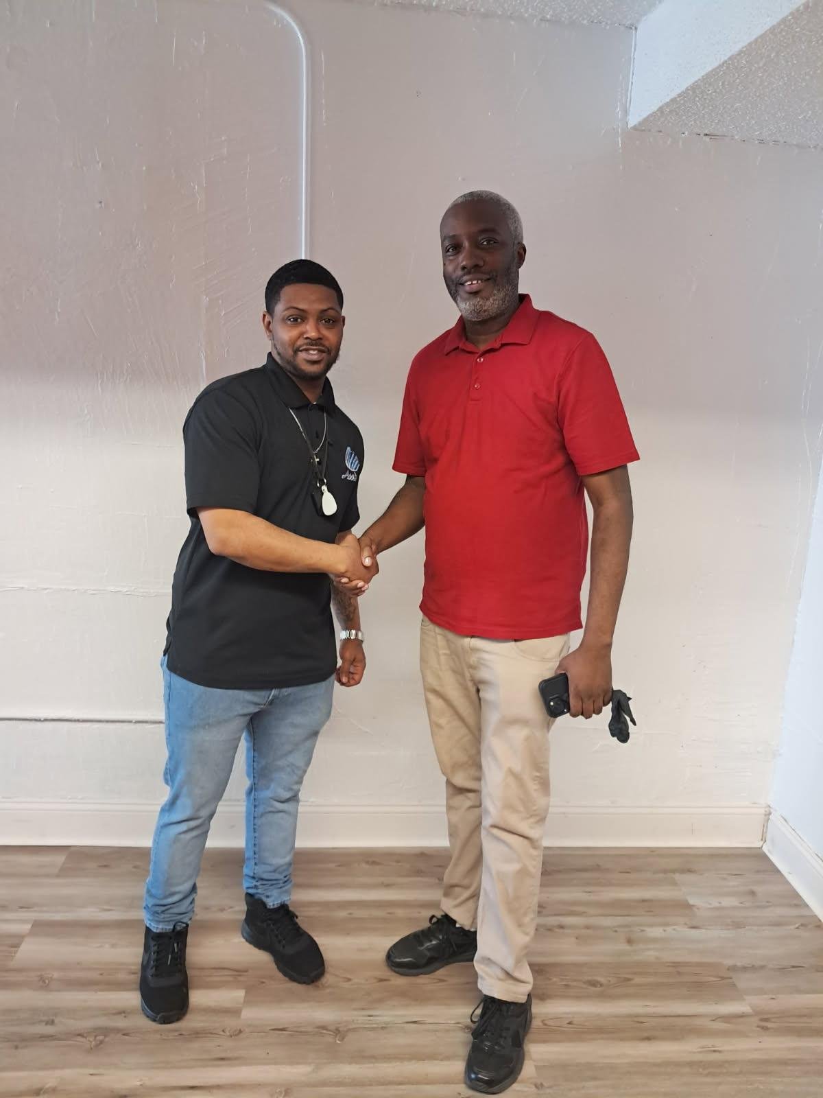 Two men shaking hands indoors, one wearing a black polo shirt and jeans, the other wearing a red polo shirt and khakis, smiling at the camera.