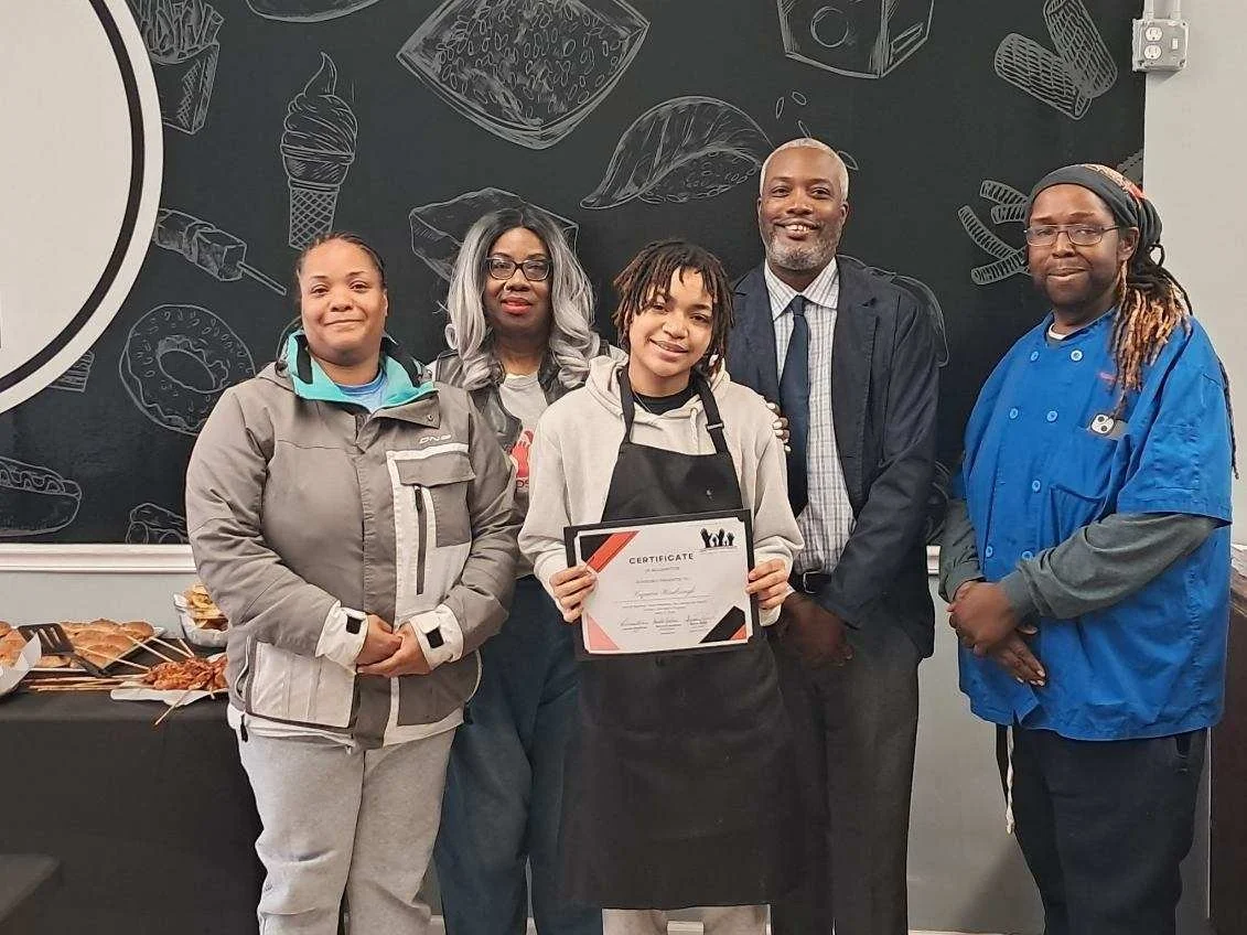 Group of five people, including a young person holding a certificate, standing in front of a chalkboard with food illustrations and a table of food, possibly at an awards or graduation event.
