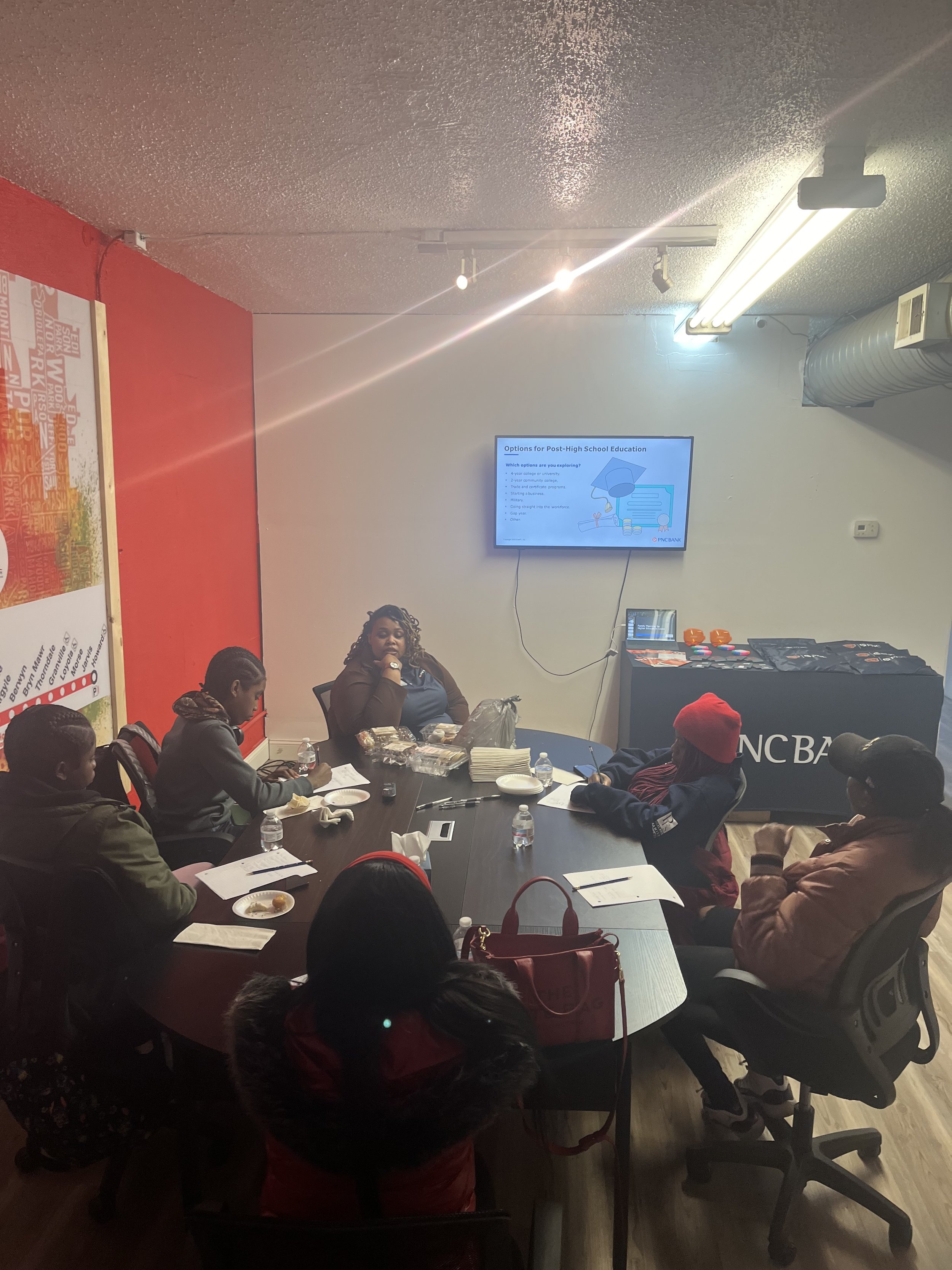 Group of people sitting around a conference table in a room, attending a presentation displayed on a wall-mounted TV screen.