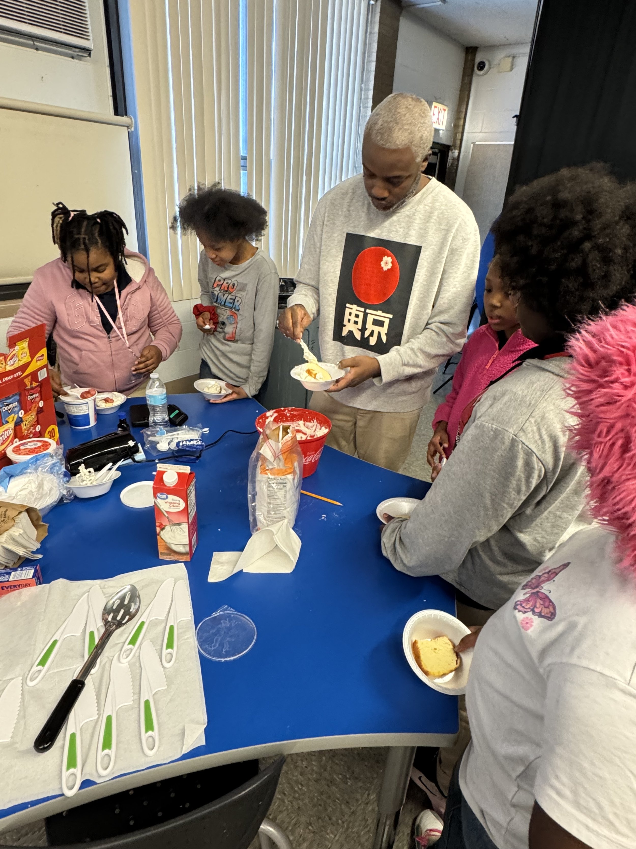 A group of children and an adult gathered around a blue table, serving and eating dessert. The table has utensils, food supplies, and a red container, with vertical blinds and an exit sign visible in the background.