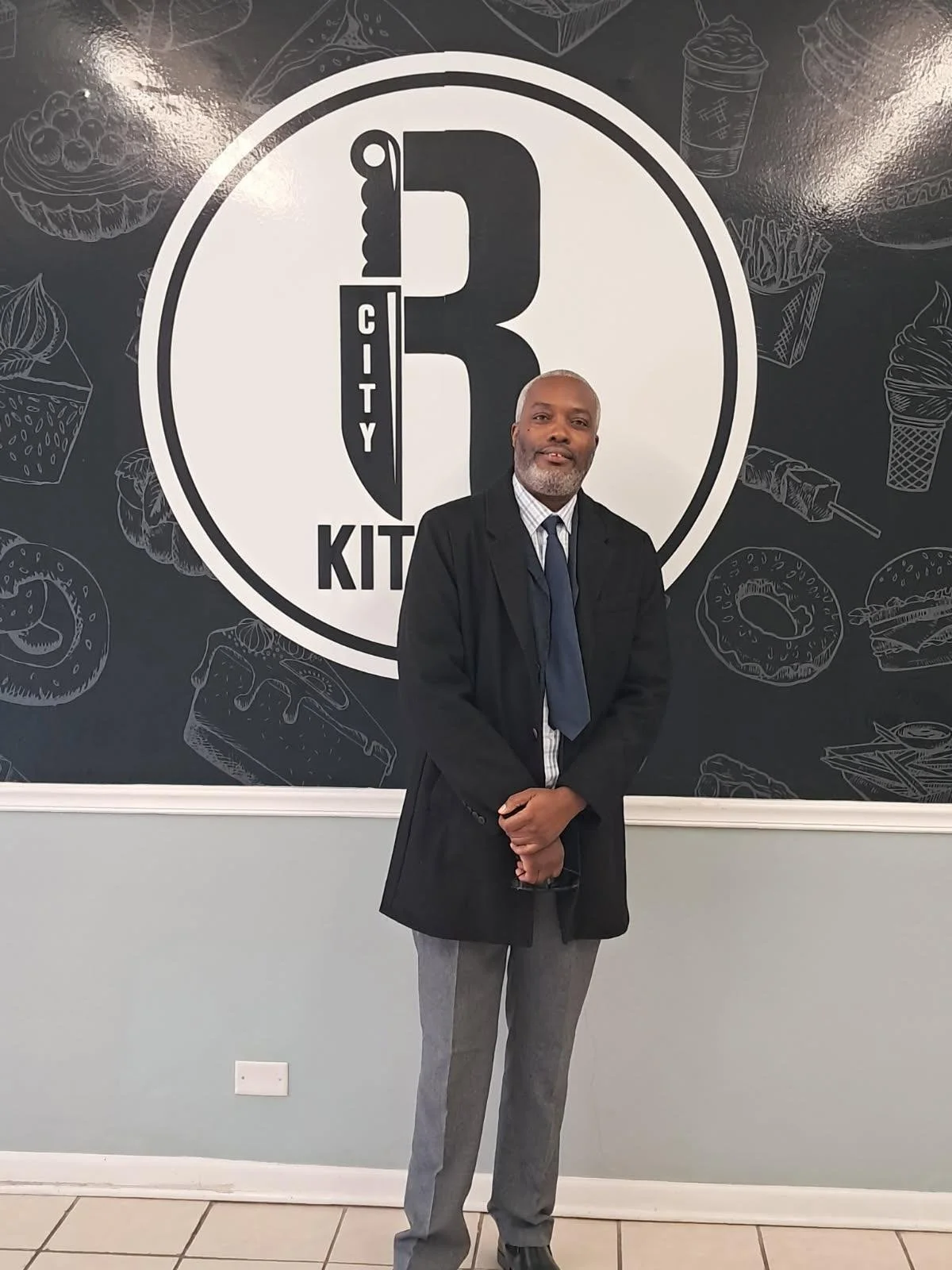 A man in a suit and coat standing inside a restaurant with a large logo on the wall behind him that says "City R Kitchen" with a spoon and knife forming the letter R. The background features illustrations of various desserts and fast food items.