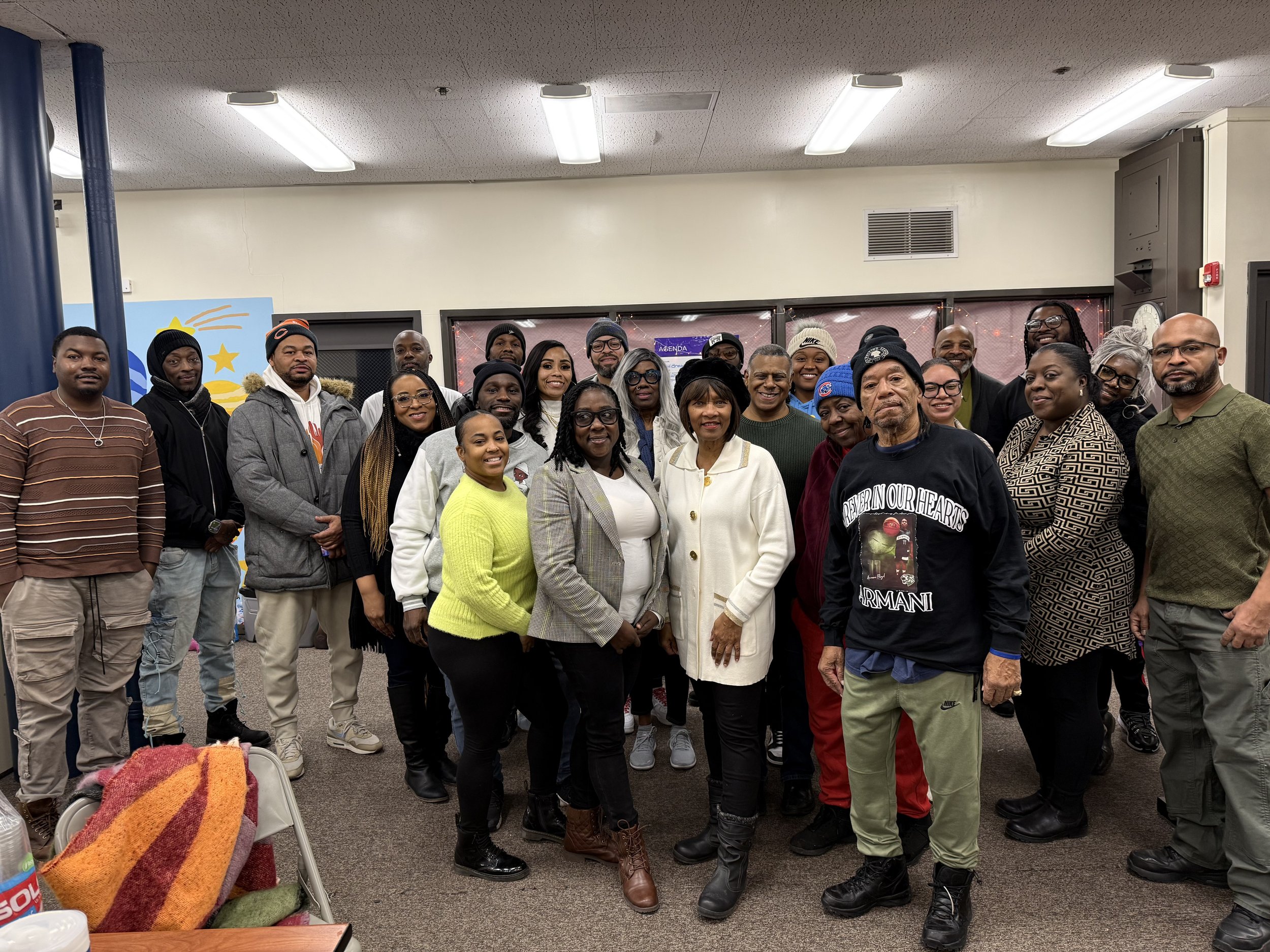 Group of diverse adults gathered indoors, smiling for a photo, in a room with colorful wall decorations and overhead fluorescent lighting.