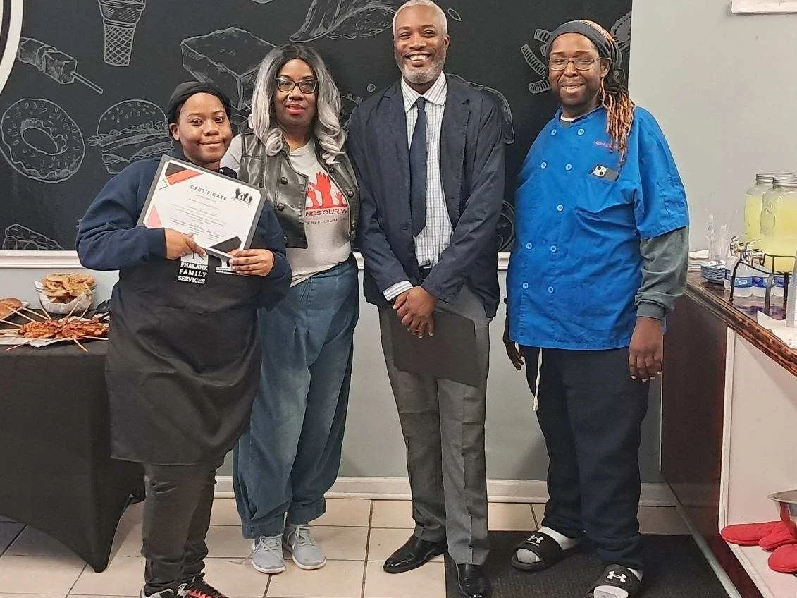 Group of four individuals standing indoors in front of a chalkboard wall with food-themed drawings. The person on the far left is holding a certificate and wearing a black apron, the second person from the left has gray hair, glasses, and casual clot