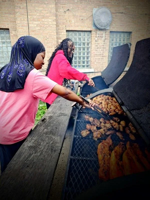 Two women cooking chicken on an outdoor grill. One woman in a pink shirt and navy headscarf, the other in a pink hoodie and glasses, are tending to the chicken on the barbecue.
