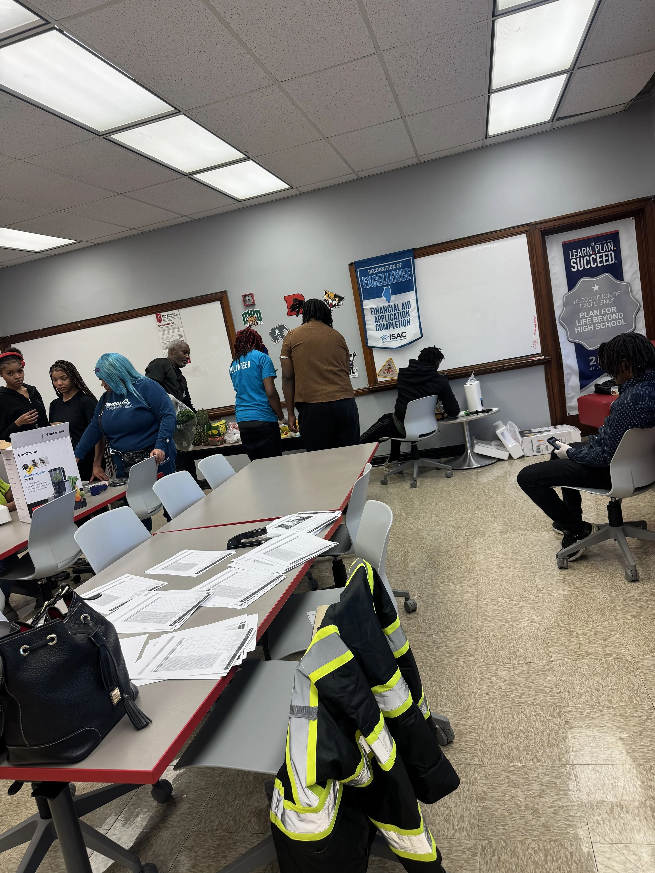 A room with tables, chairs, and people. Papers and a backpack on the tables. Several individuals stand near a whiteboard with blue and gray posters about excellence and planning. A person in safety gear hangs on the back of the chair. Others are seat