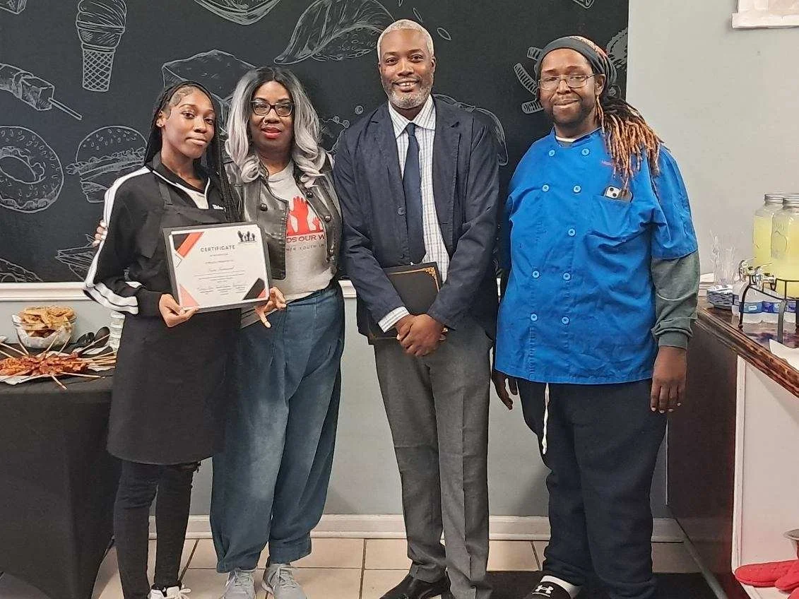 Four individuals standing in a room, with a chalkboard wall featuring drawings of food items like donuts and sandwiches, and a table with food and drinks on the side. The first person is holding a certificate. The group appears to be celebrating or p