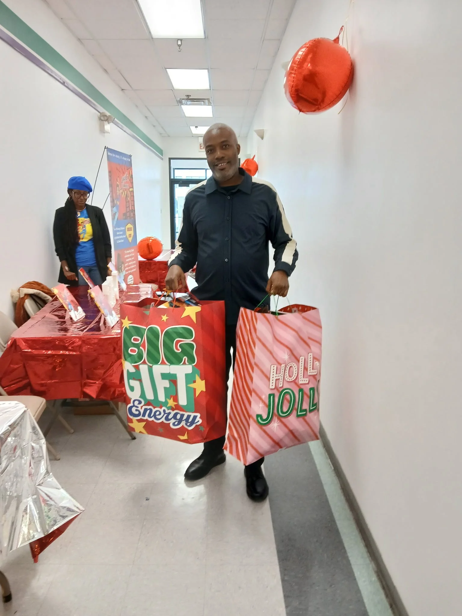 A man smiling and holding two large festive shopping bags, one with 'Big Gift Energy' written on it and the other with 'Holly Jolly' written on it, standing in a room decorated for Christmas.