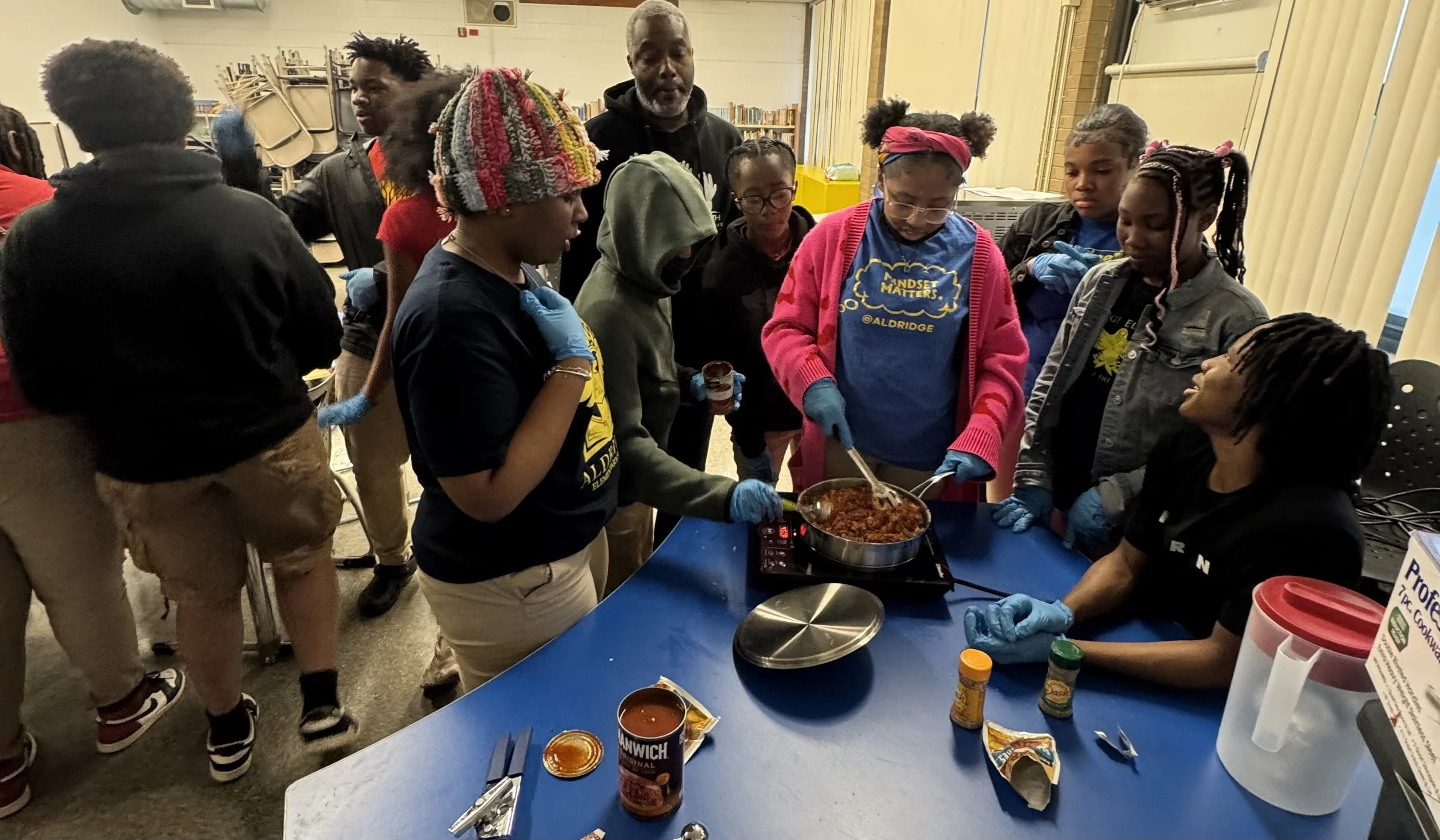A group of children and a woman in a classroom preparing and serving a meal, with the woman cooking chili or stew on a portable stove, while the children observe and participate.