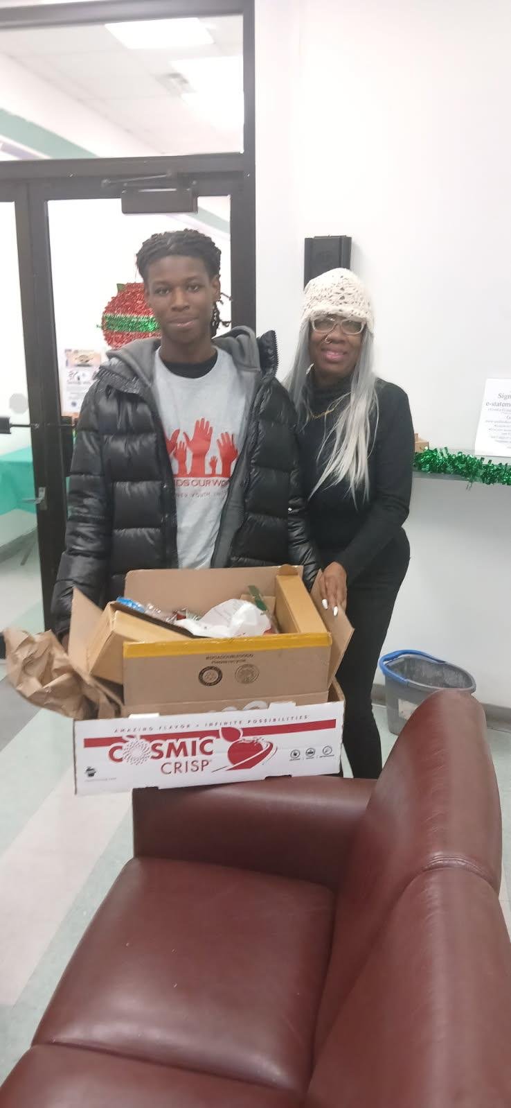 A young man and an older woman standing behind a table with a box of food in a room decorated with holiday ornaments. The young man is wearing a grey t-shirt and a black puffer jacket, and the woman is wearing glasses, a white knitted hat, and a blac
