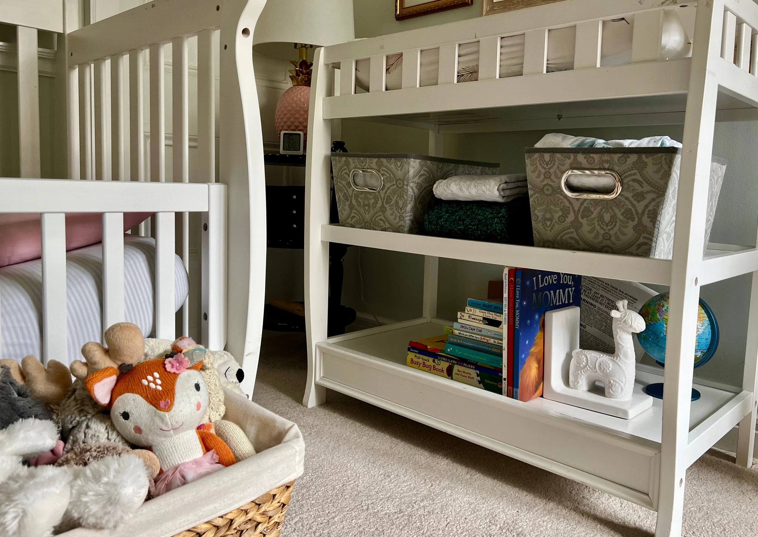 A nursery room with white cribs, a wicker basket filled with plush toys including a stuffed fox, shelves with books, a small globe, and decorative storage bins.