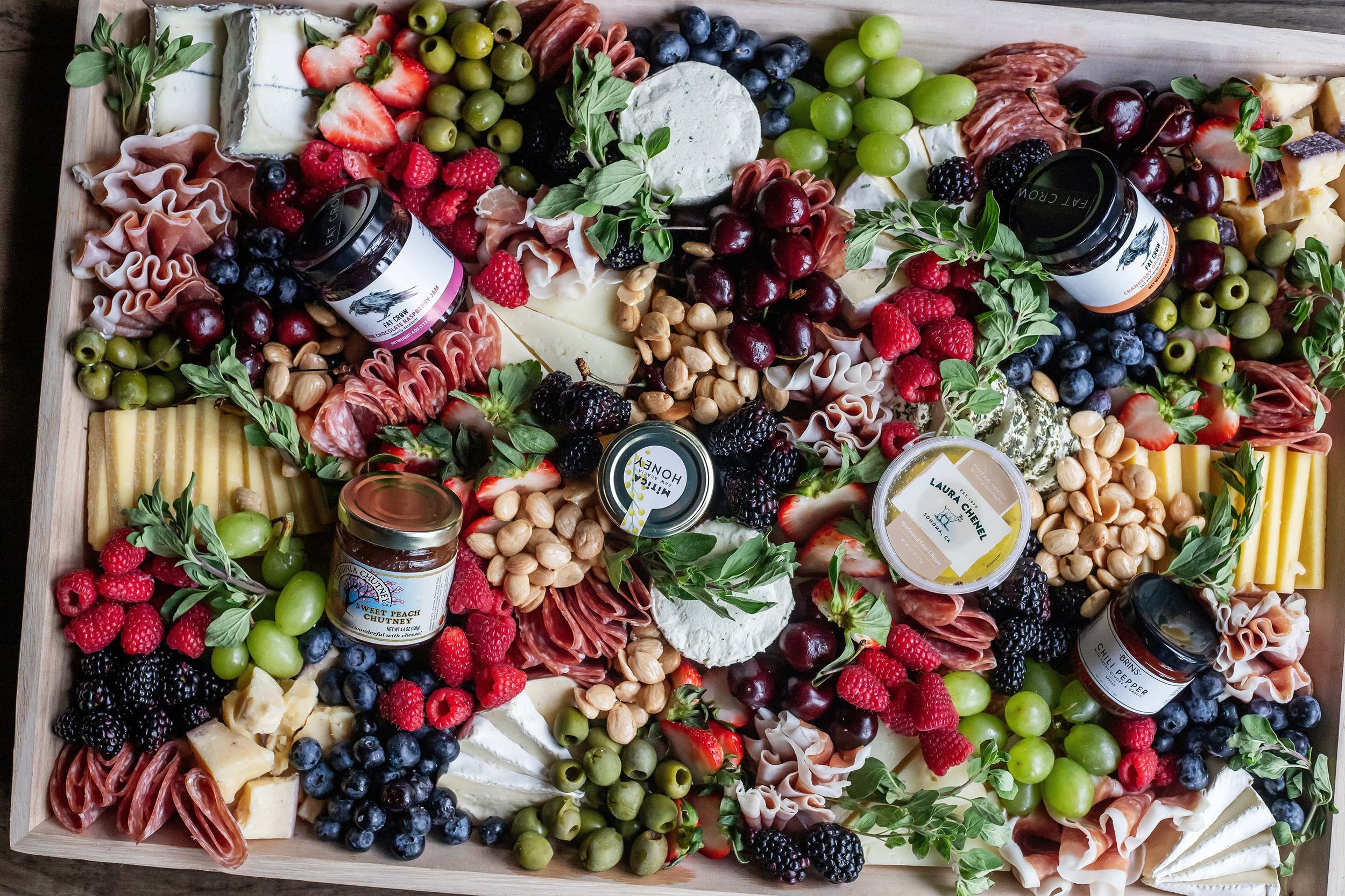 A large charcuterie board featuring assorted cheeses, grapes, berries, nuts, cured meats, fresh herbs, and jars of spreads and chutneys.