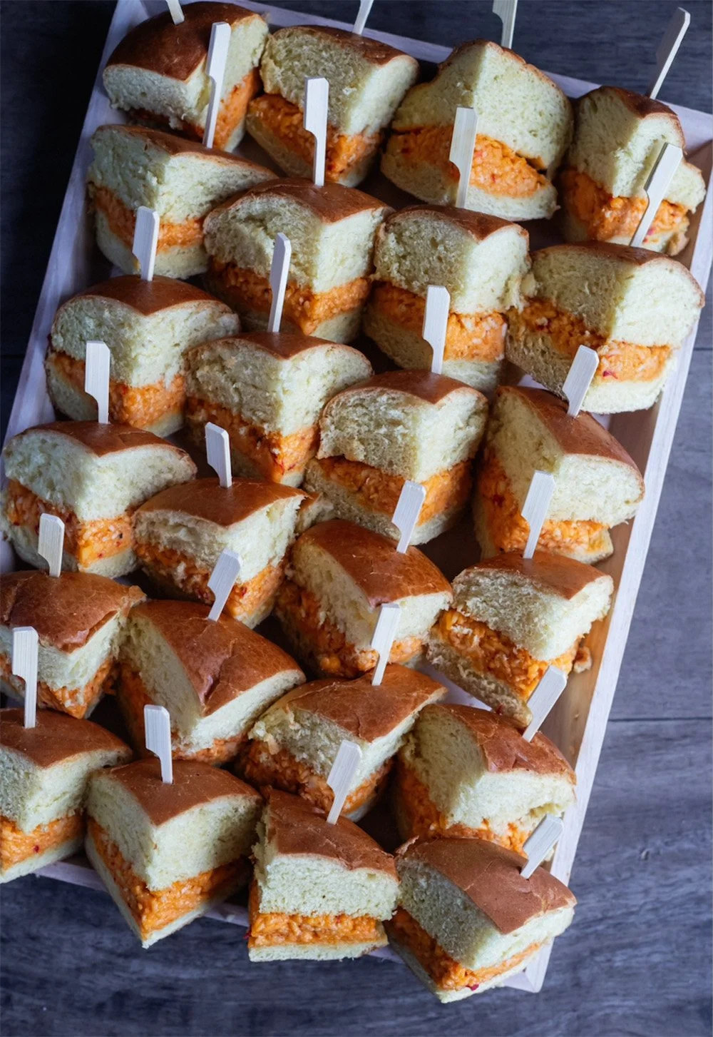 A tray of mini sandwiches with white bread and orange filling; some sandwiches have pickles, garnished with small white flags.